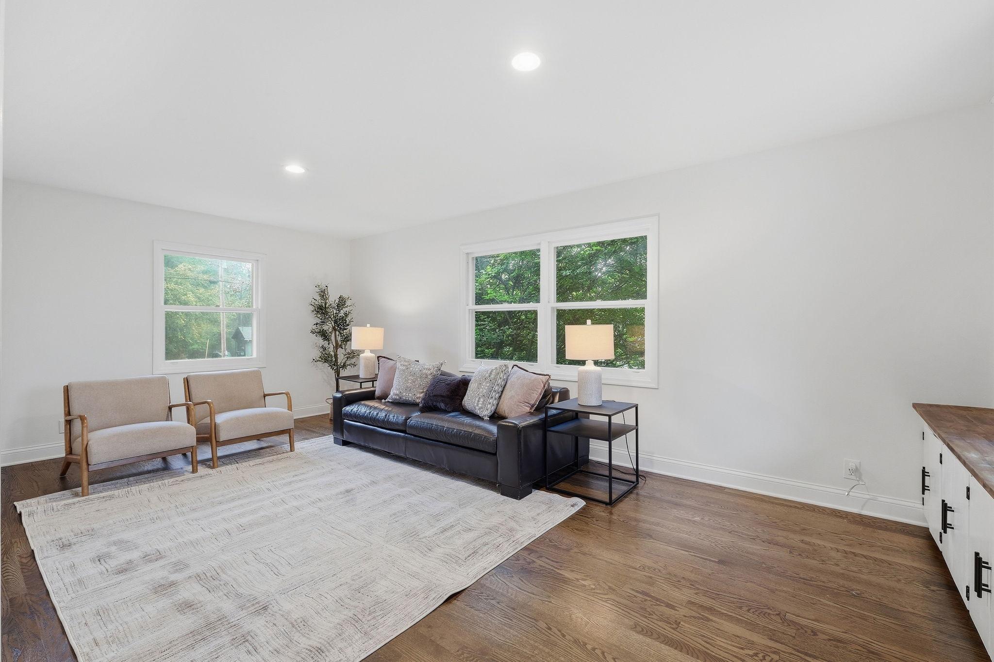 Living room with a black leather sofa, beige armchairs, hardwood floors, and leafy views through two windows.