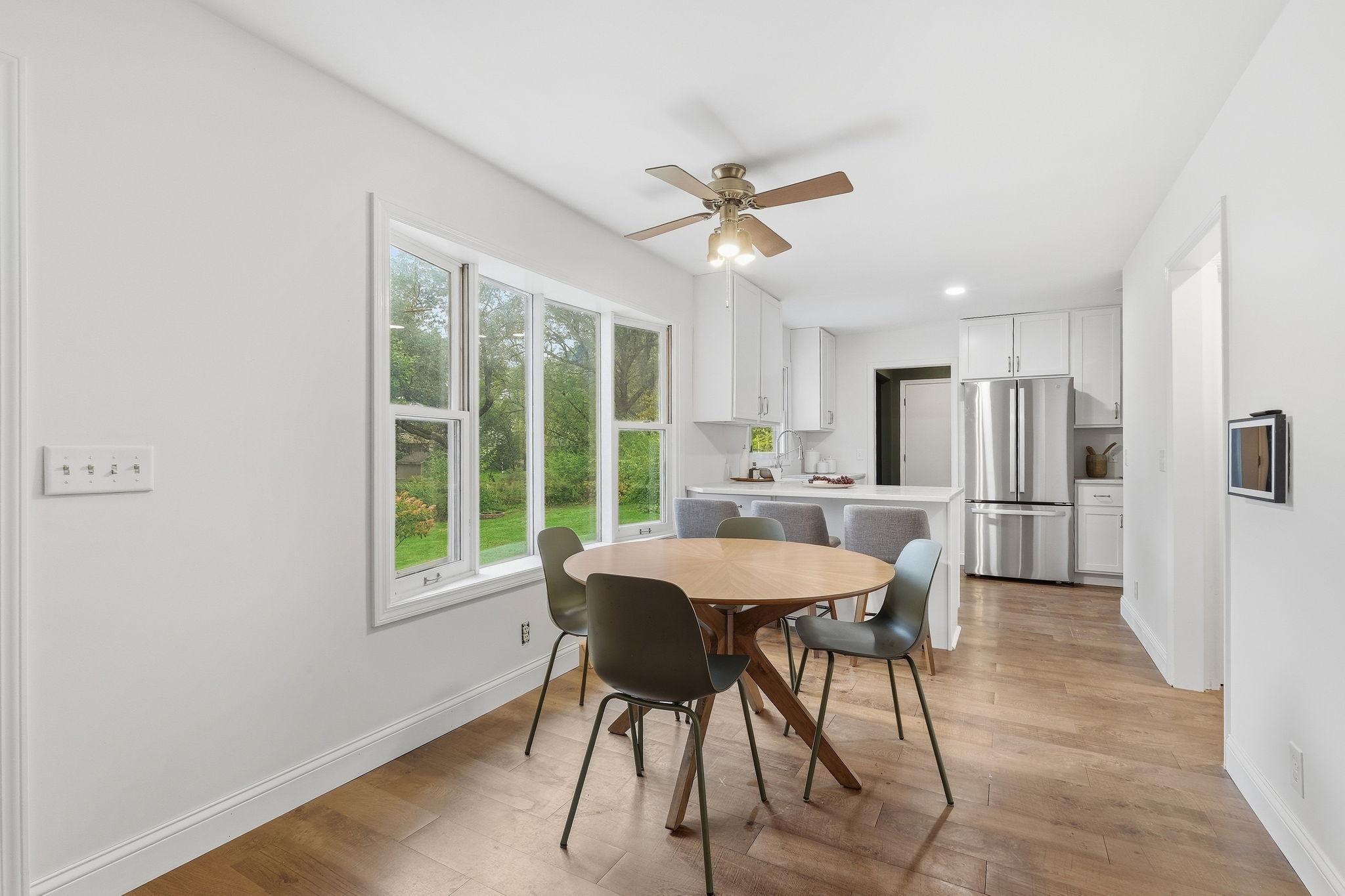 Dining area with a round wooden table beside large windows, opening into a bright kitchen.