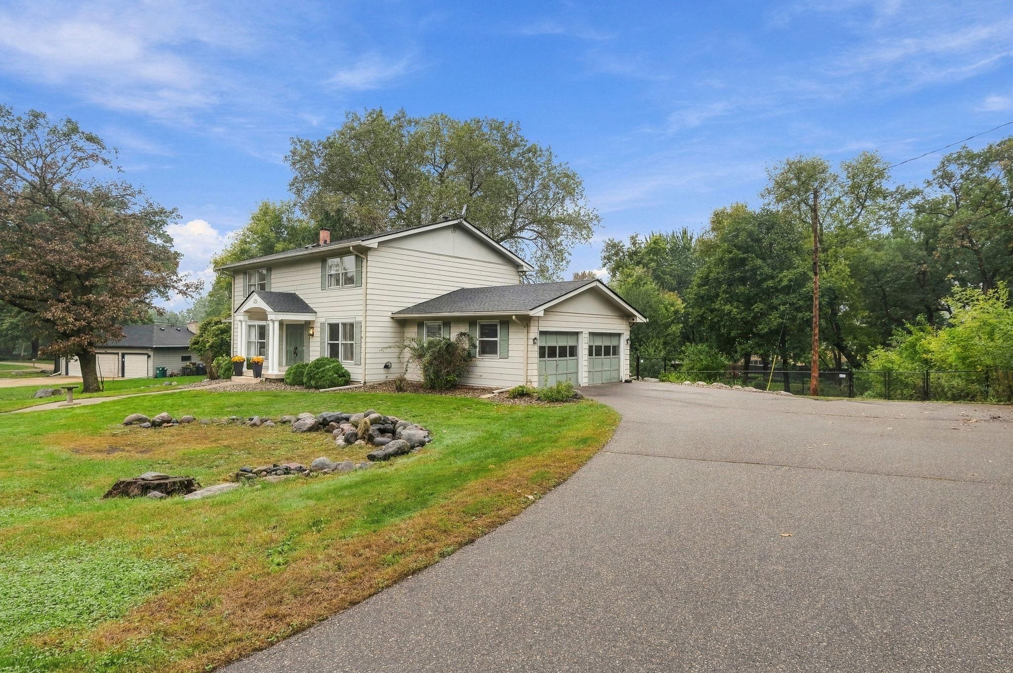 Two-story light-sided house with an attached two-car garage and broad driveway, framed by mature trees.