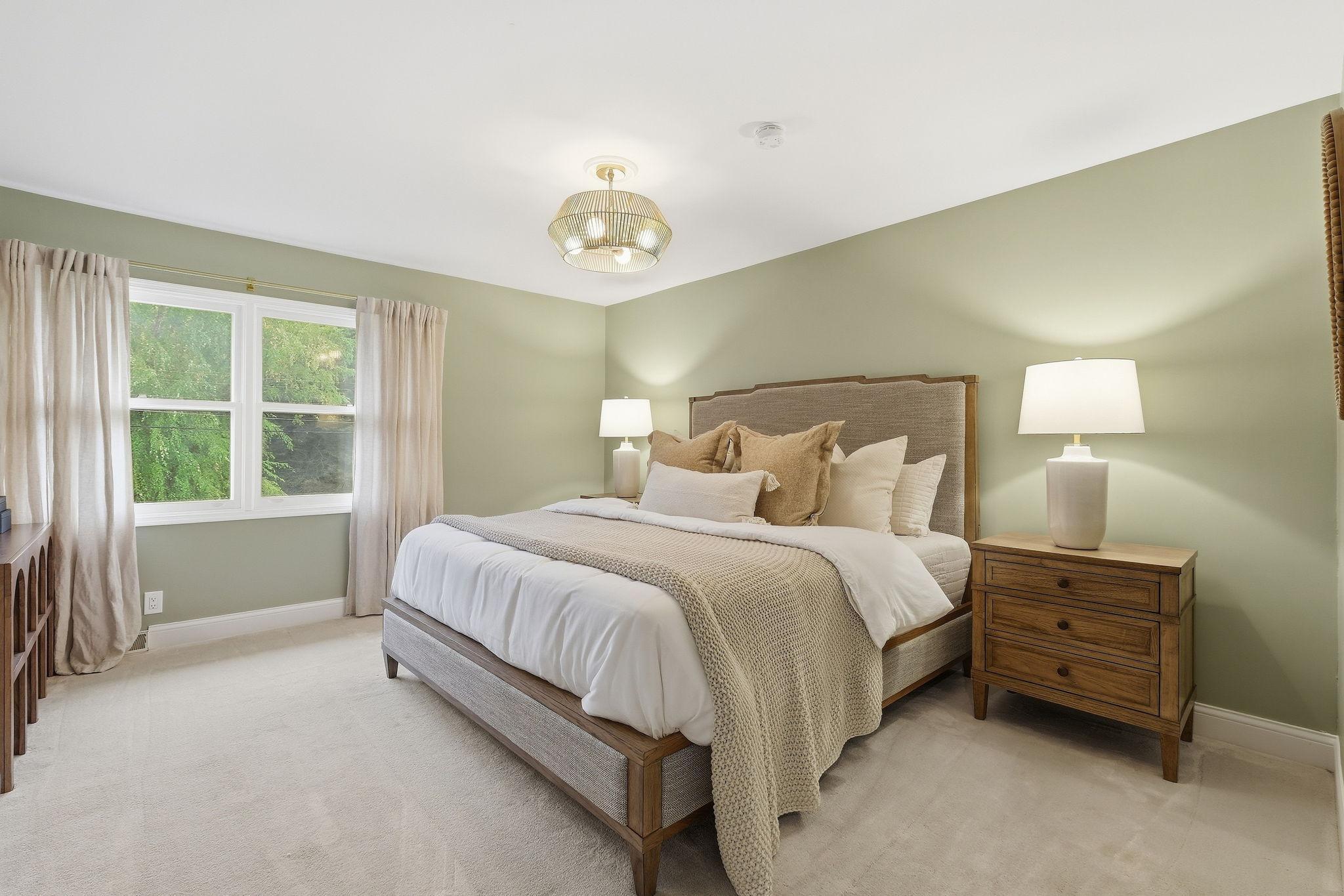 Bedroom with a wood-framed bed, neutral layered bedding, sage-green walls, and a large curtained window.