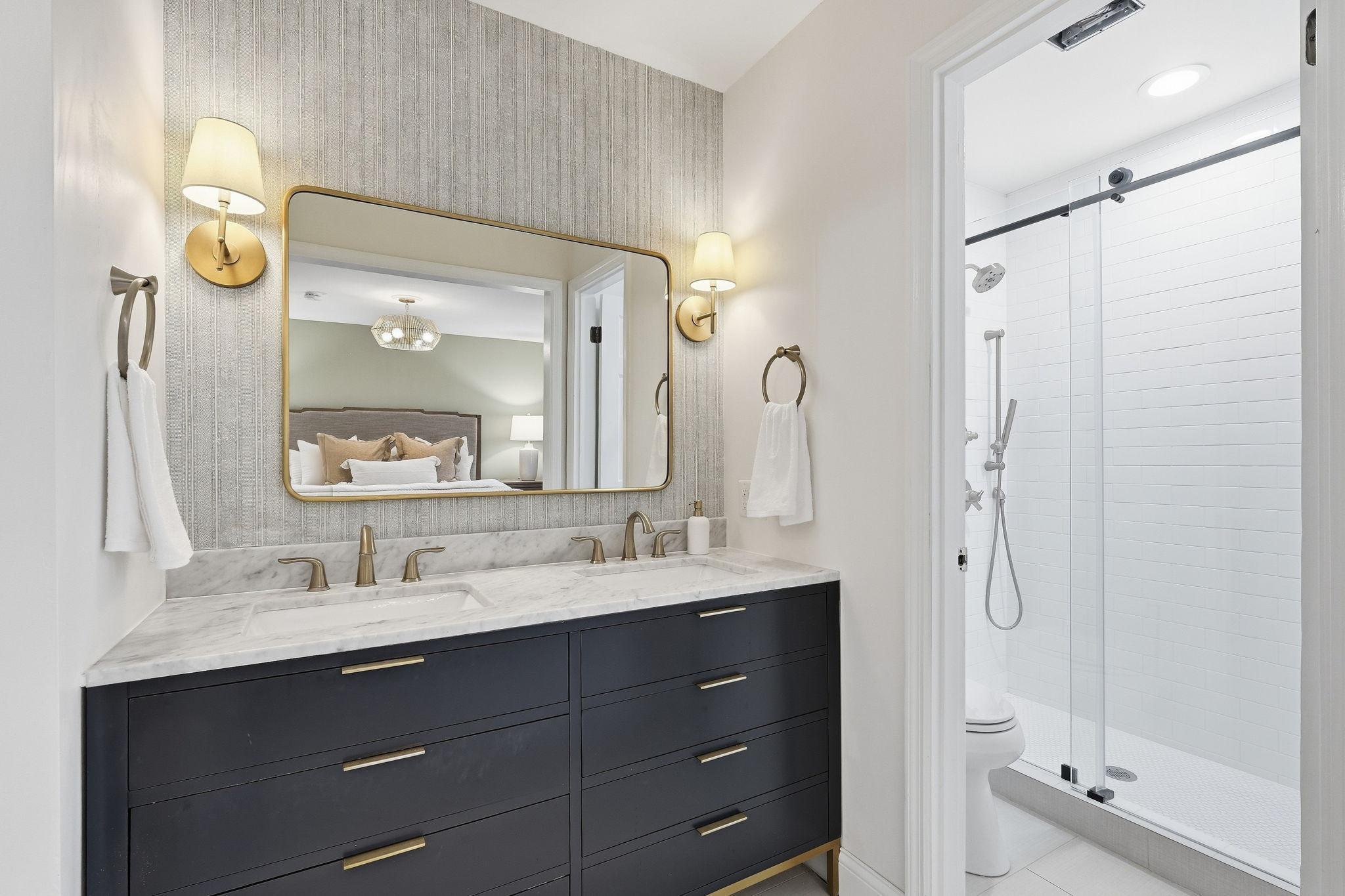 Bathroom with dark double vanity, marble countertop, brass fixtures, and glass-enclosed white tile shower.