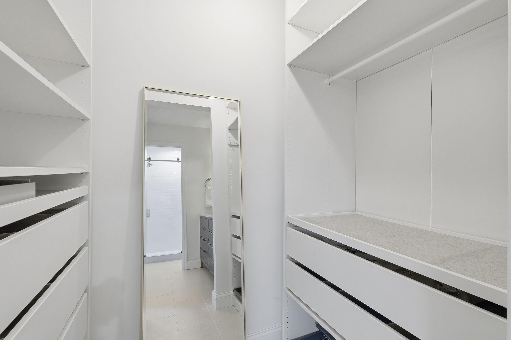 Walk-in closet with white built-in shelves and drawers, a full-length mirror, and an adjoining bathroom.