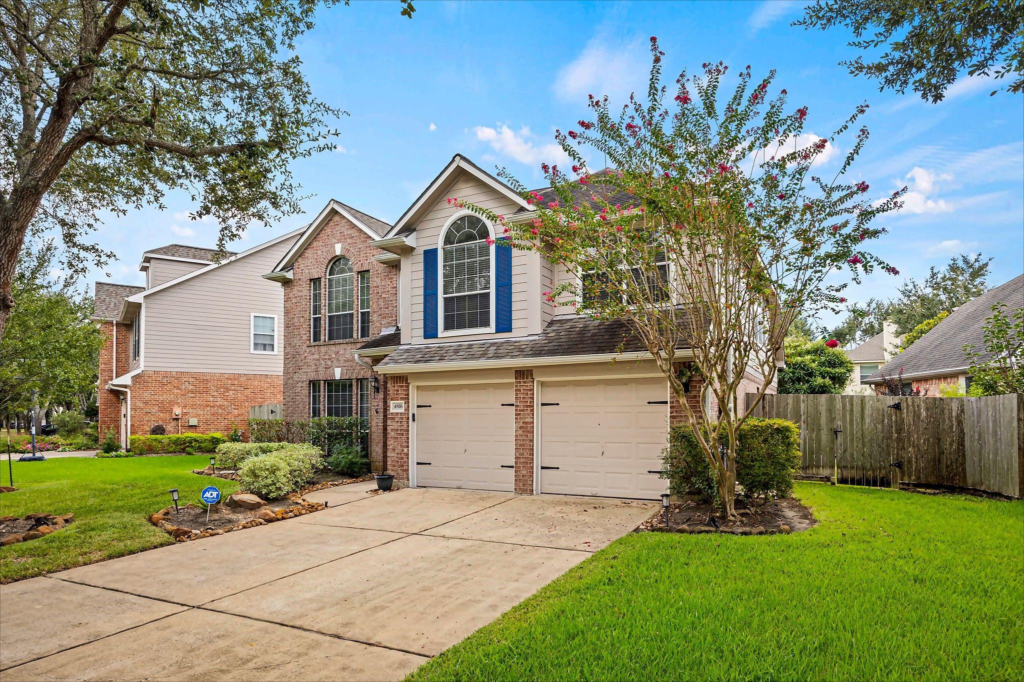 Two-story brick-and-siding home with blue shutters, a two-car garage, and flowering trees beside the driveway.