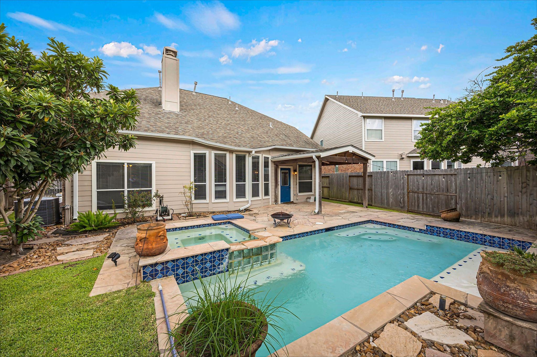 Backyard pool and raised spa beside a beige house, with a stone patio and covered entry.