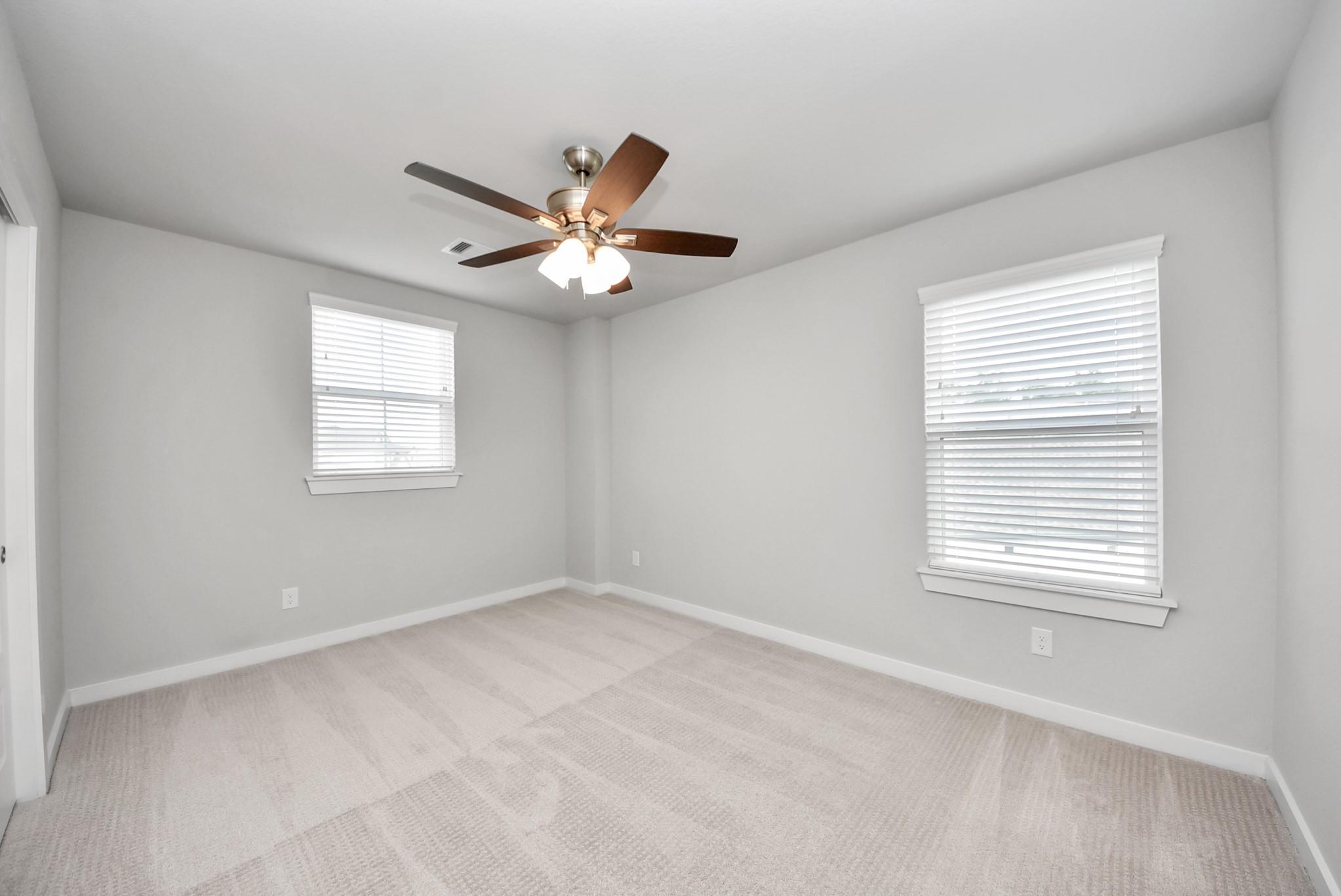 Empty carpeted room with two blind-covered windows and a ceiling fan with lights.