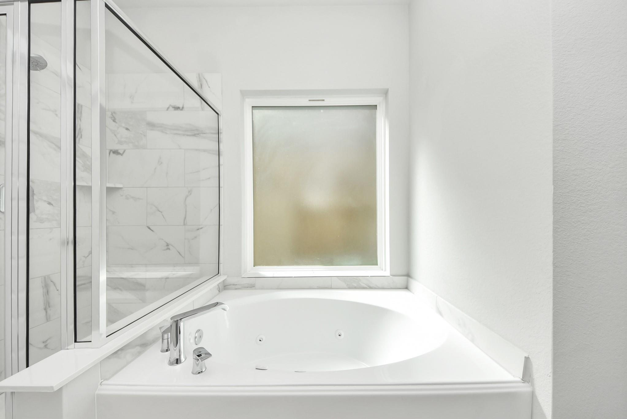 Bathroom with a white jetted tub, frosted window, and glass-enclosed marble-look tile shower.