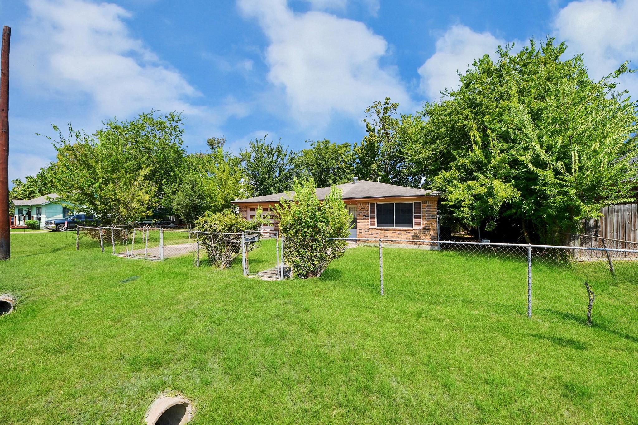 Single-story brick house behind a fenced lawn, surrounded by mature trees.