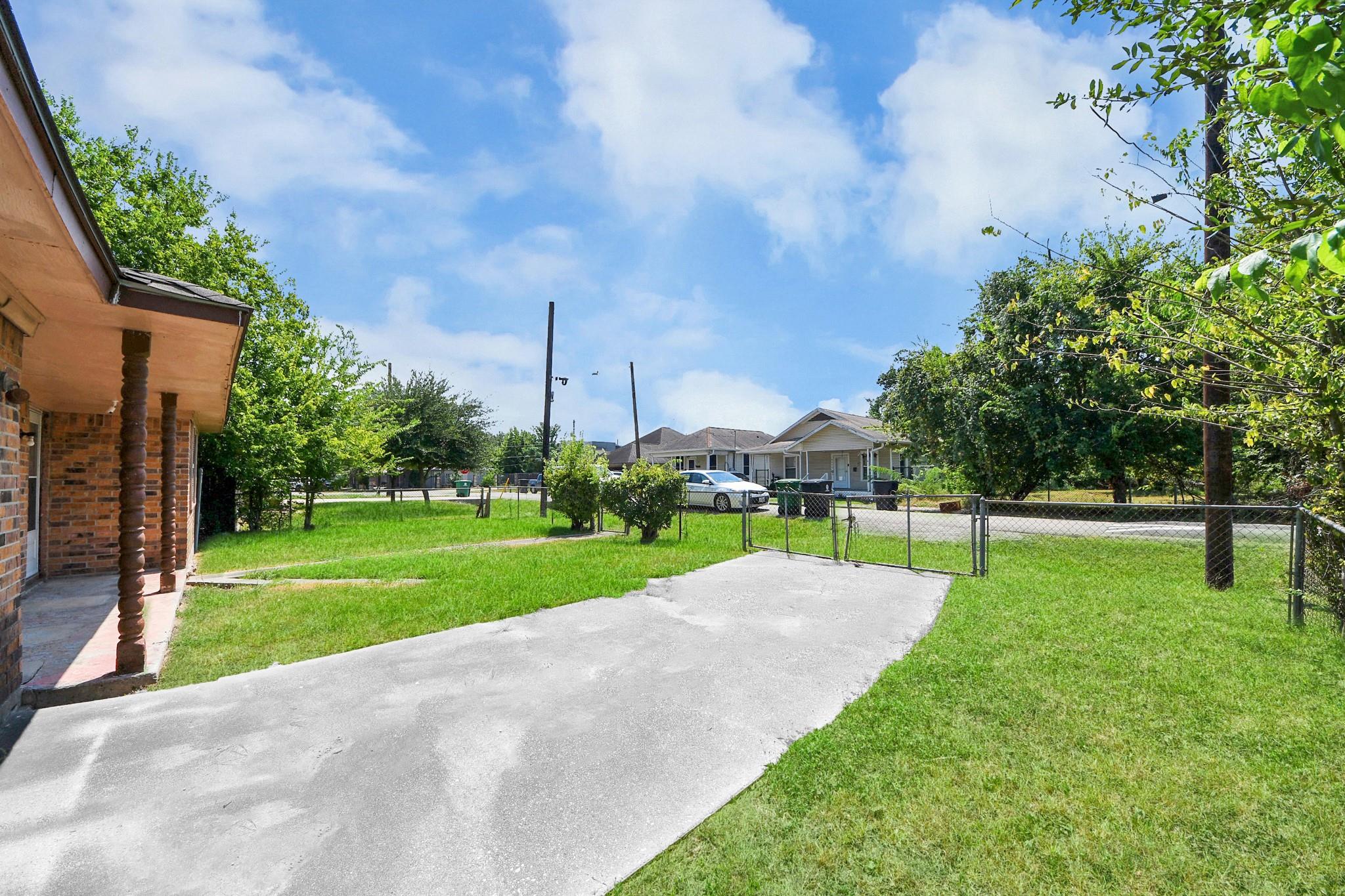 Grassy yard and concrete driveway beside a brick house, with a chain-link fence and gate.