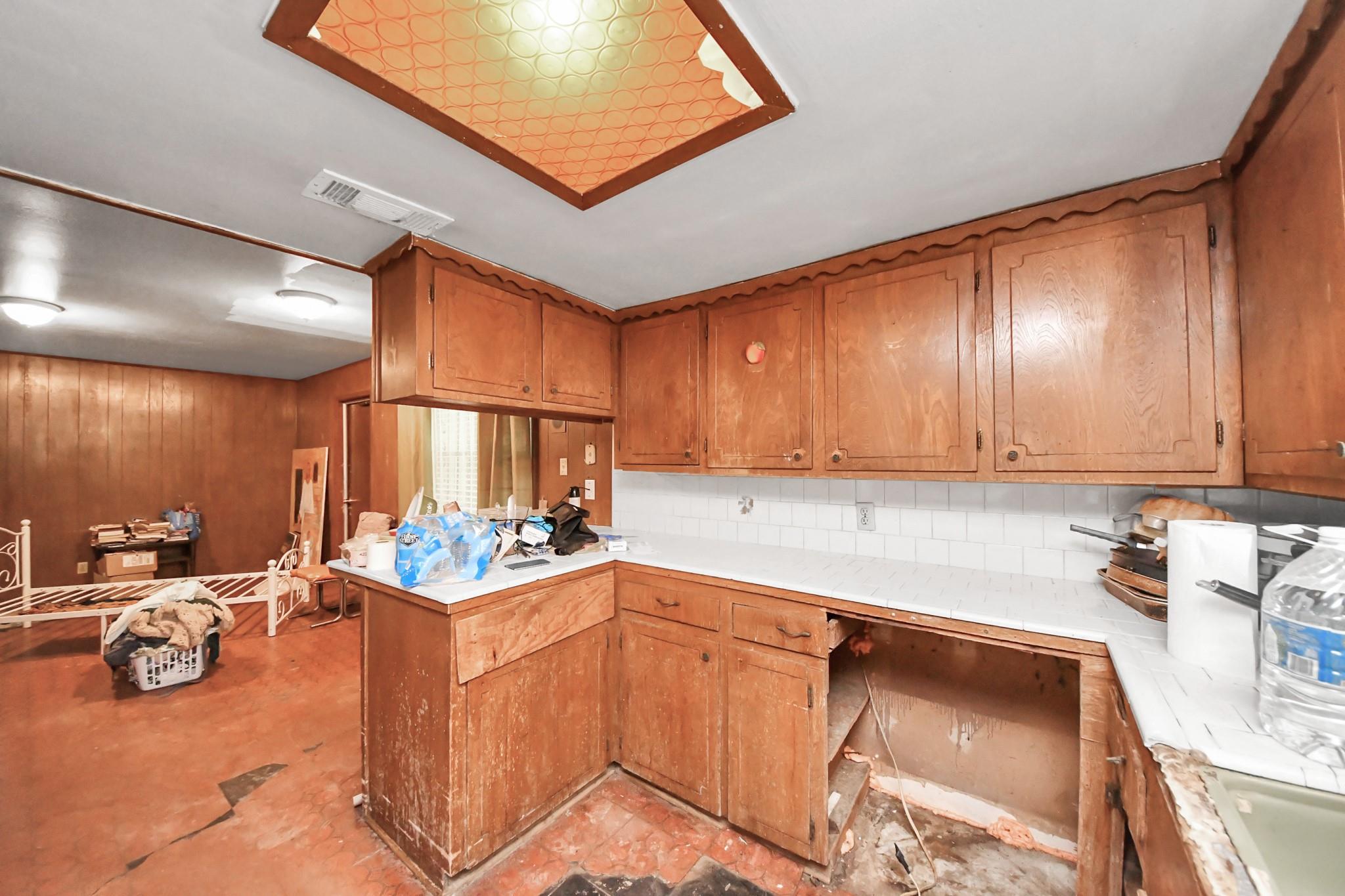 Dated kitchen with worn wood cabinets, tiled counters, and an open, unfinished section beneath the counter.