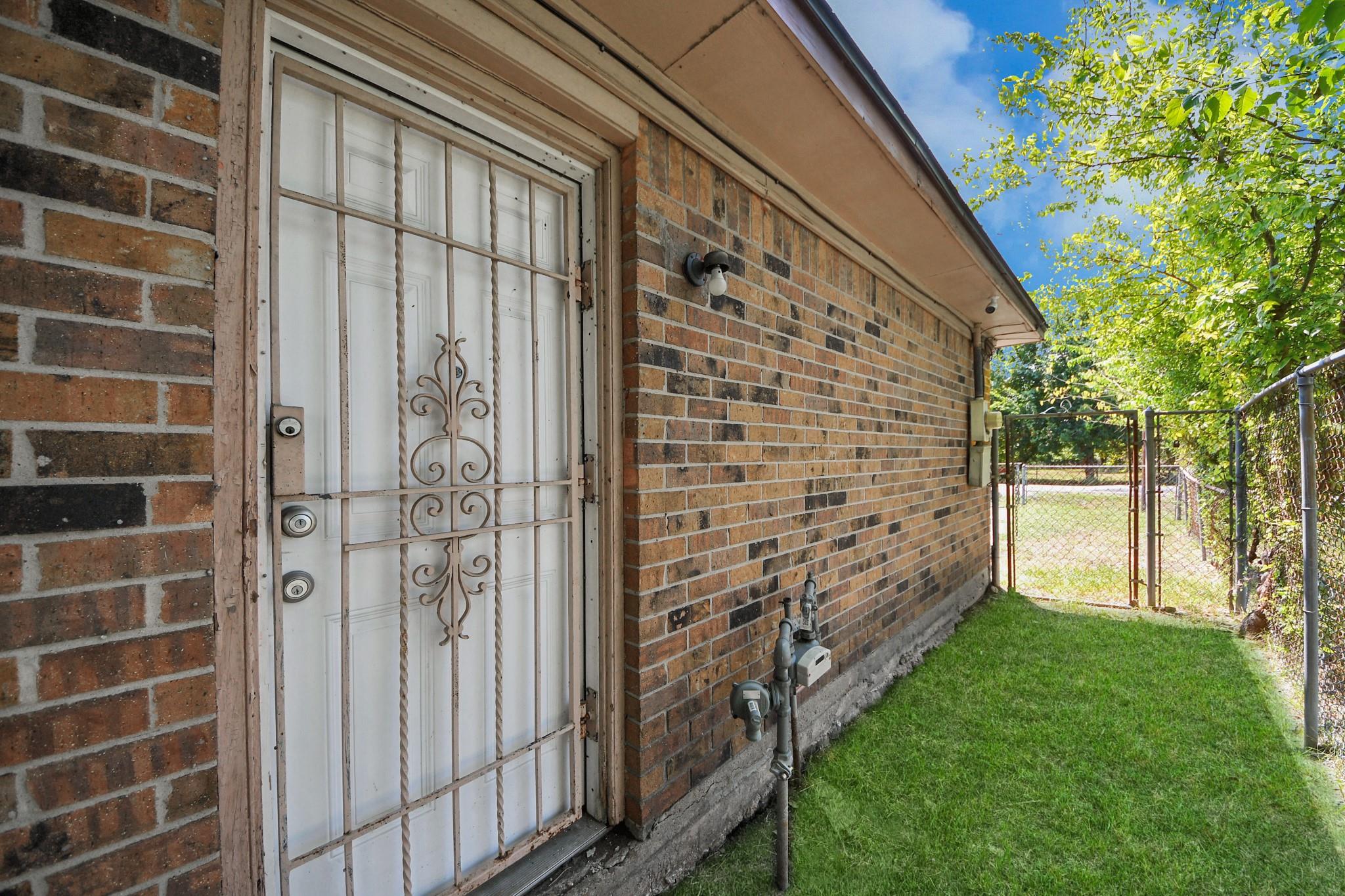 Brick house side entrance with a white security-grille door and grassy yard leading to a chain-link gate.
