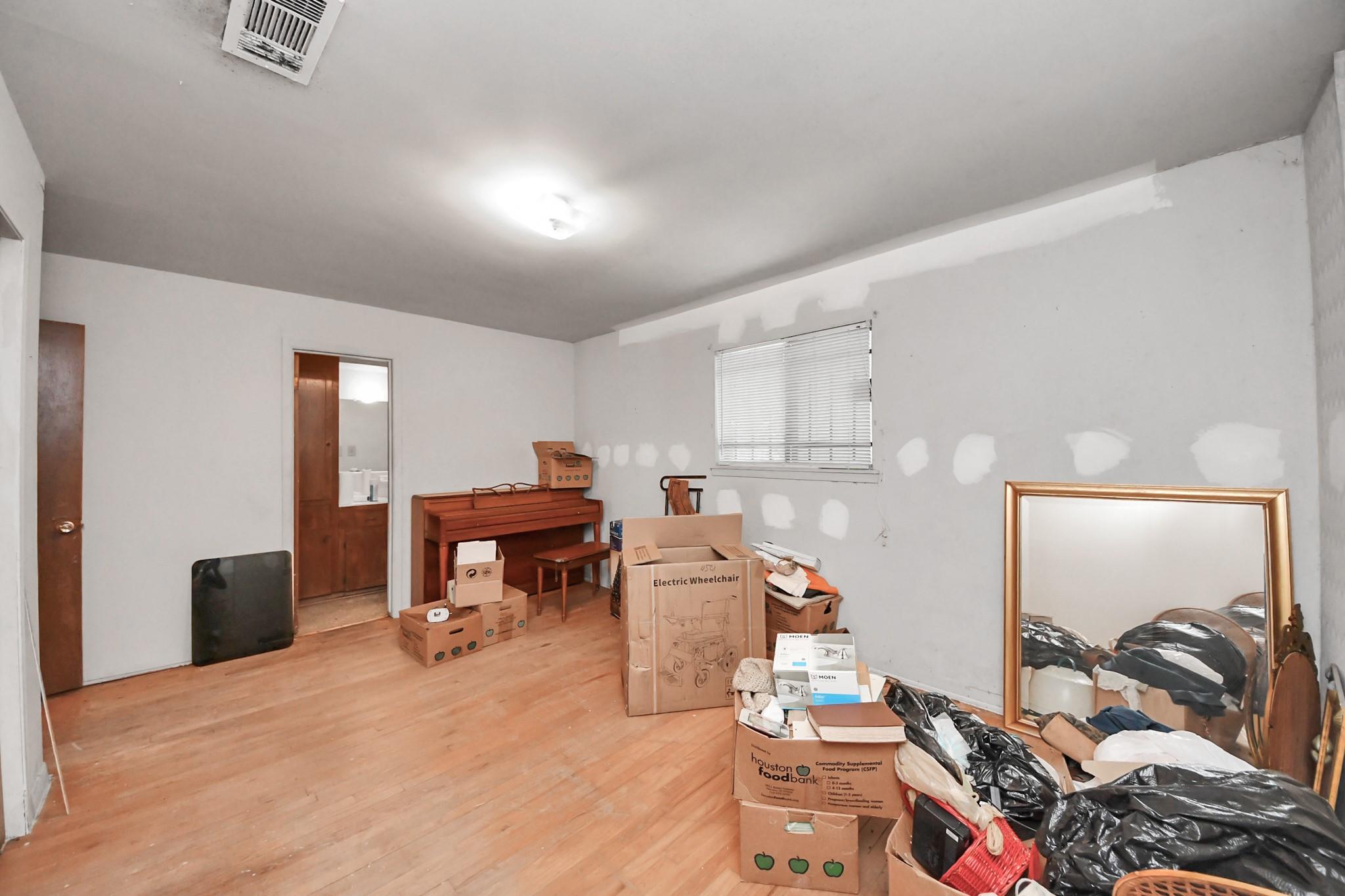 Cluttered room with a wooden piano, patched walls, closed blinds, and boxes and belongings piled beside a large mirror.
