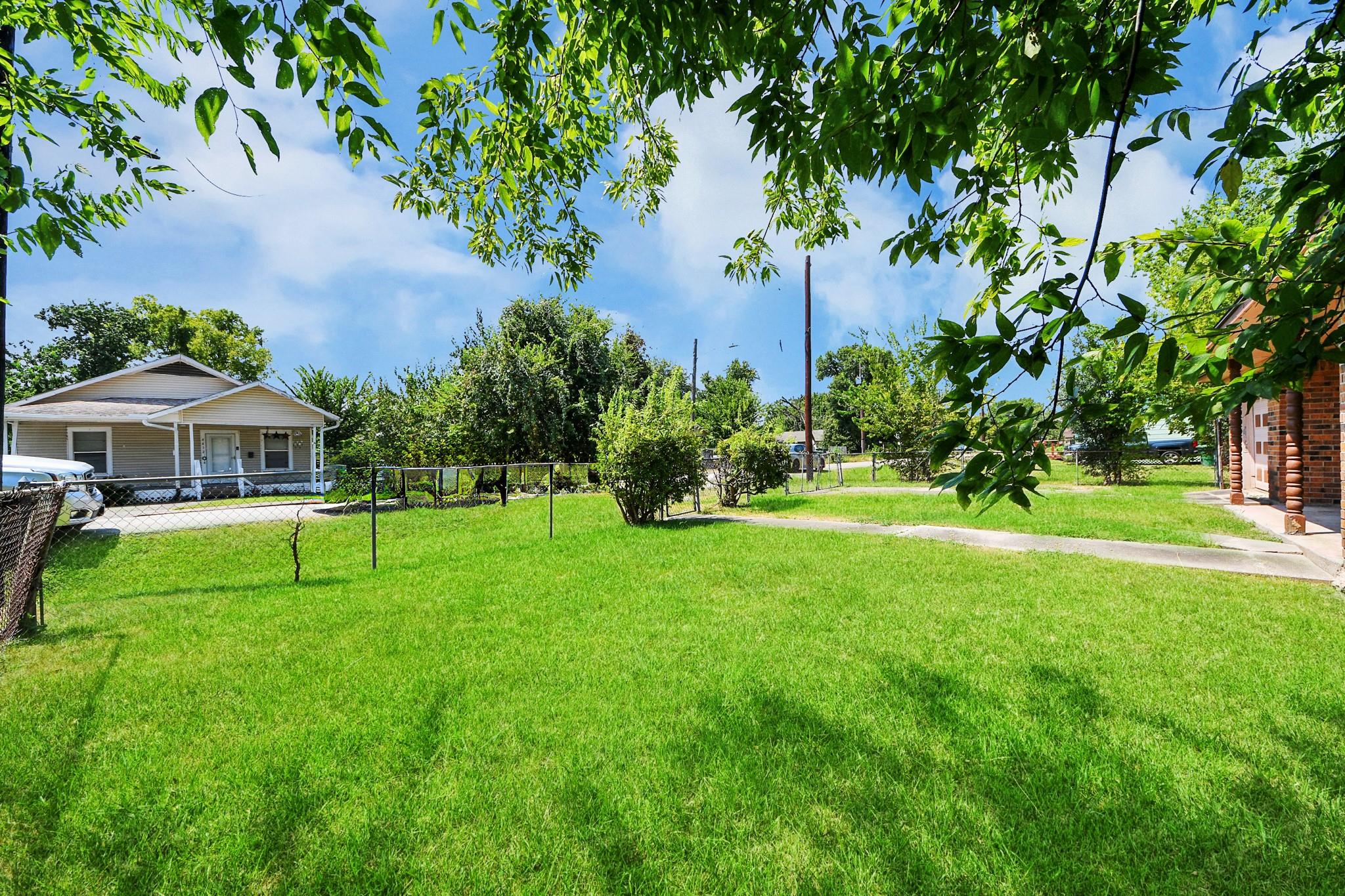 Large grassy yard with a chain-link fence, leafy branches, and part of a brick house and porch.
