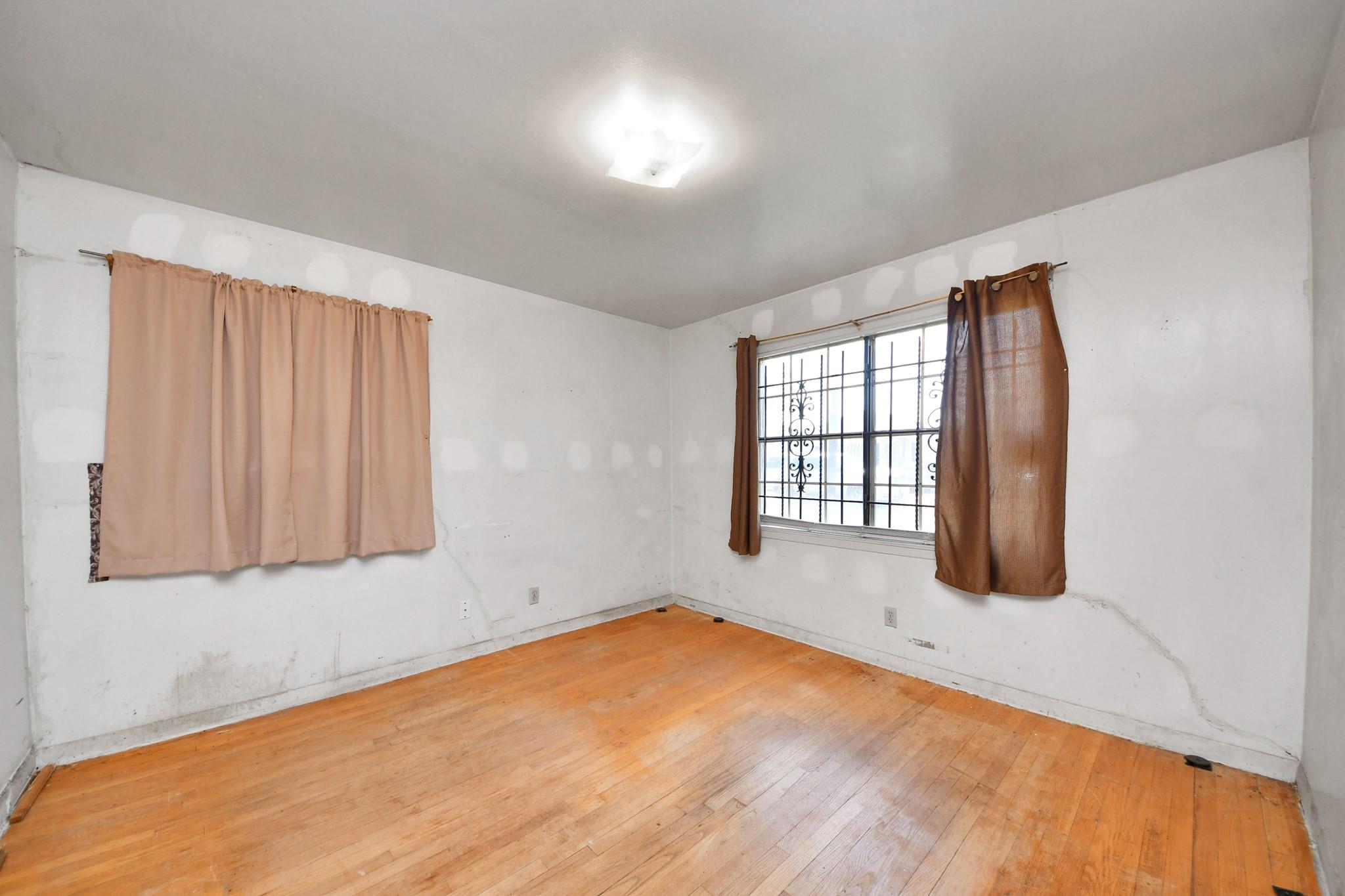 Empty room with hardwood floors, patched white walls, and curtained windows with security bars.