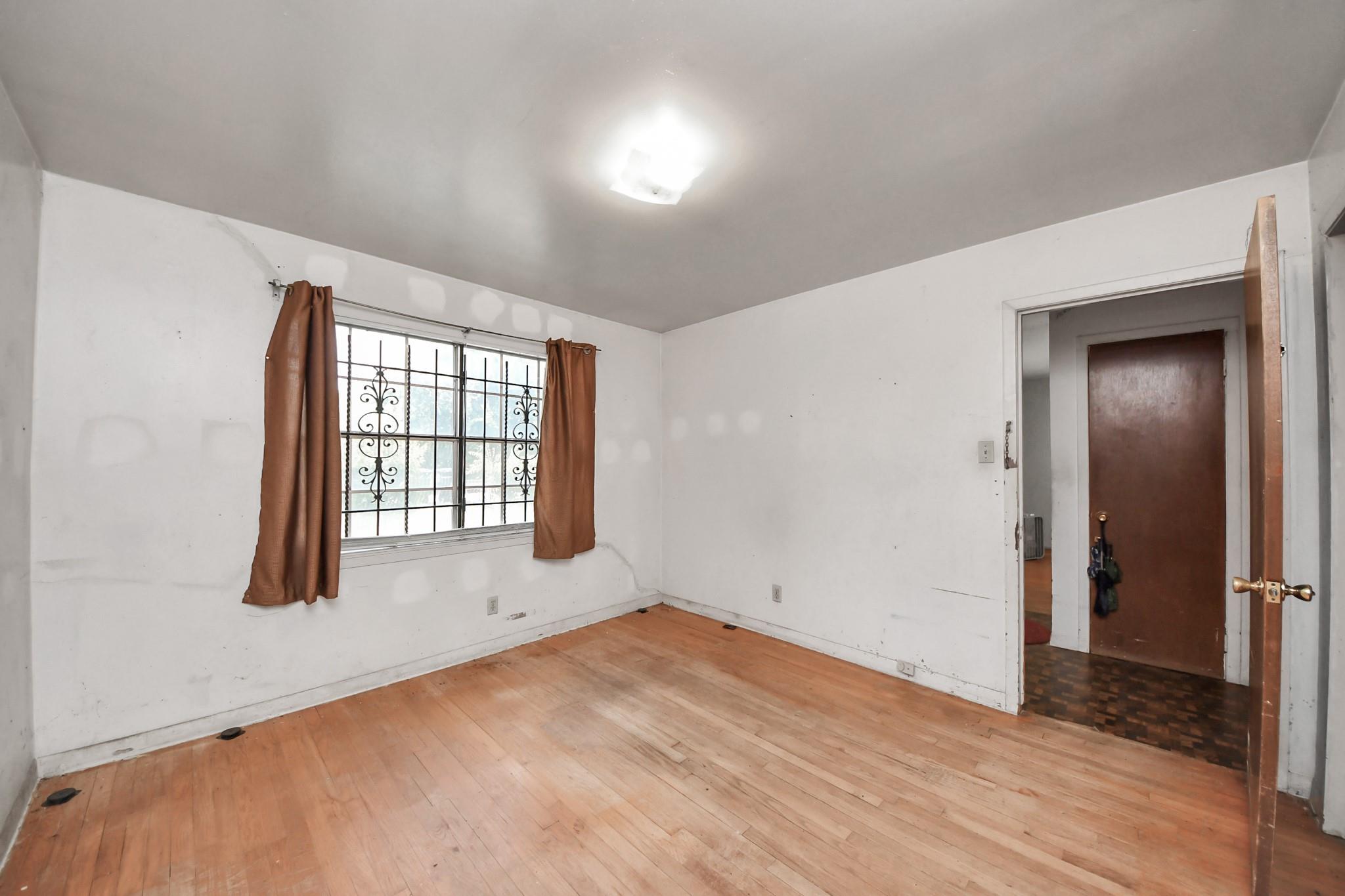 Empty room with hardwood floors, a barred window, and brown curtains.
