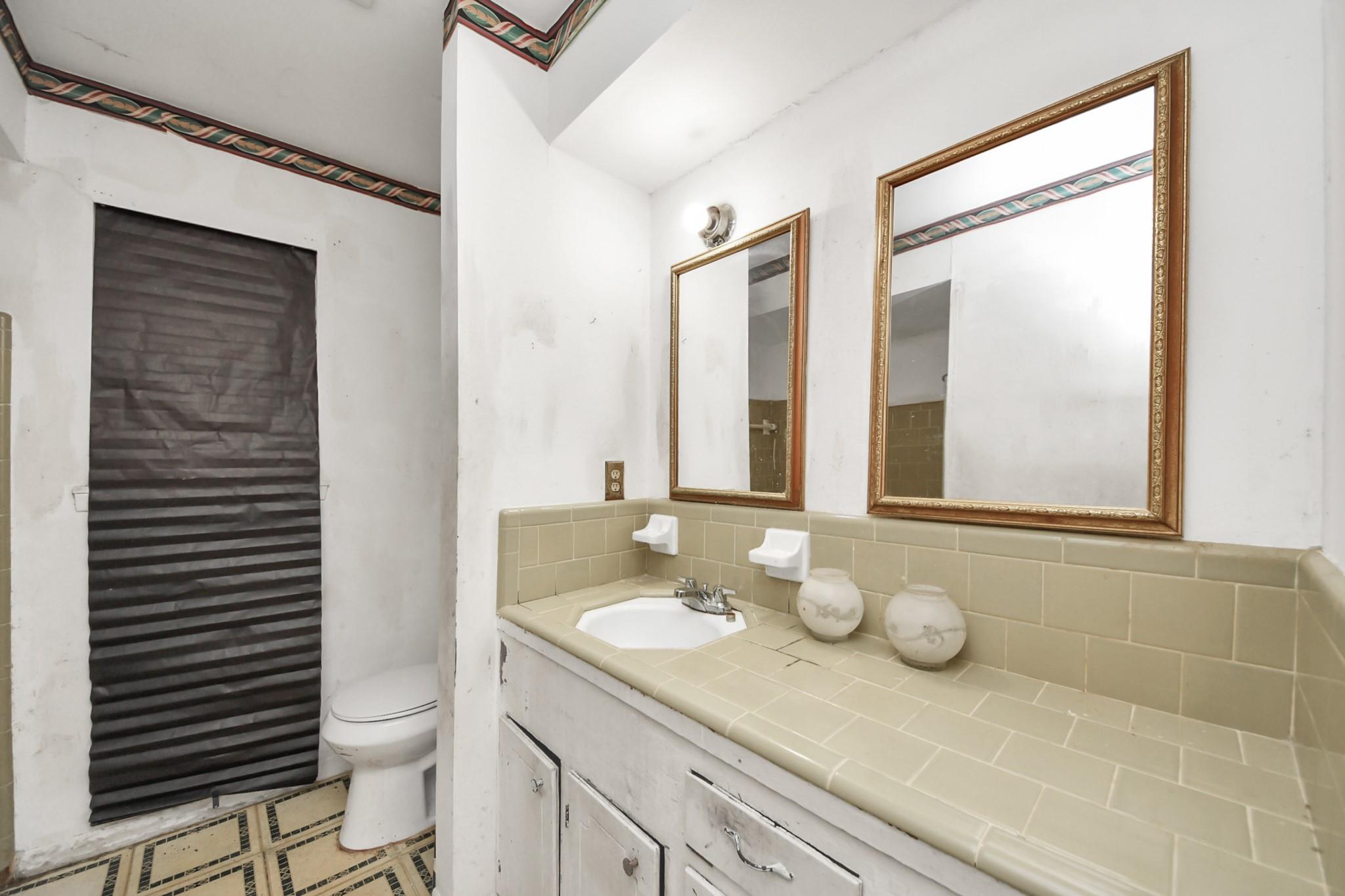 Bathroom with a beige tiled vanity, two gold-framed mirrors, and a toilet beside a shaded window.