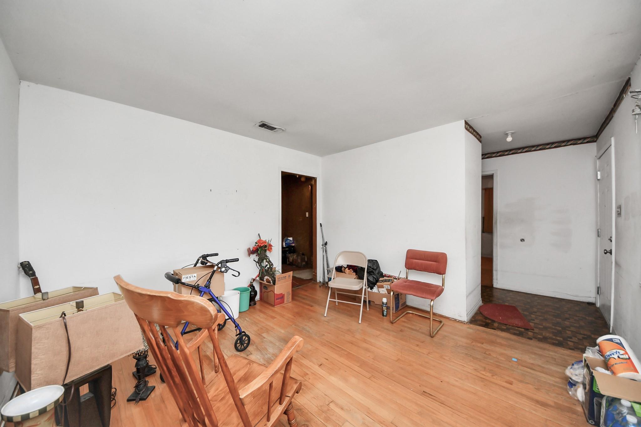 Cluttered living room with hardwood floors, scattered chairs, boxes, and an adjoining hallway.