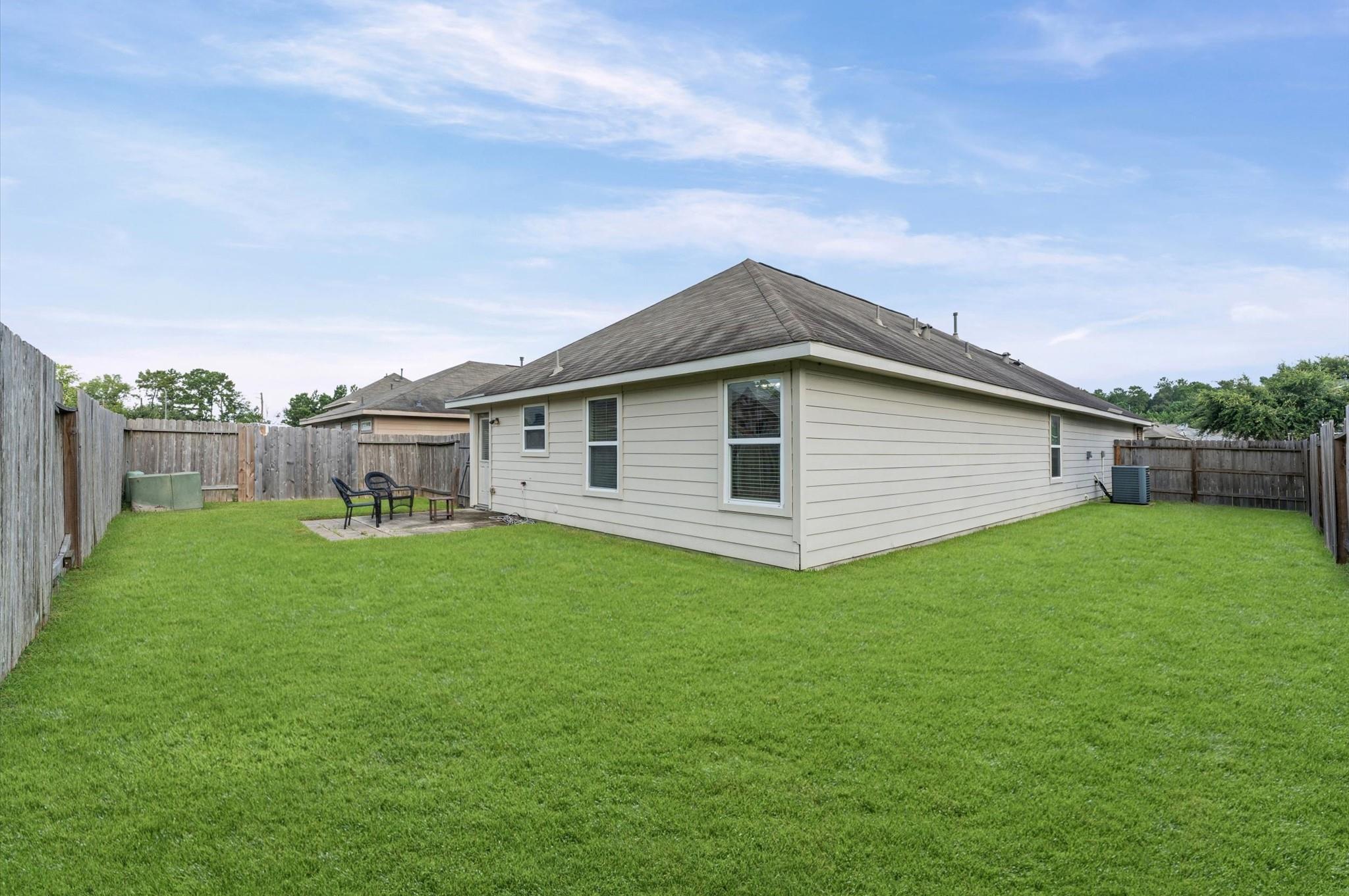 Home’s rear exterior beyond a broad fenced lawn, with a small patio and outdoor chairs.