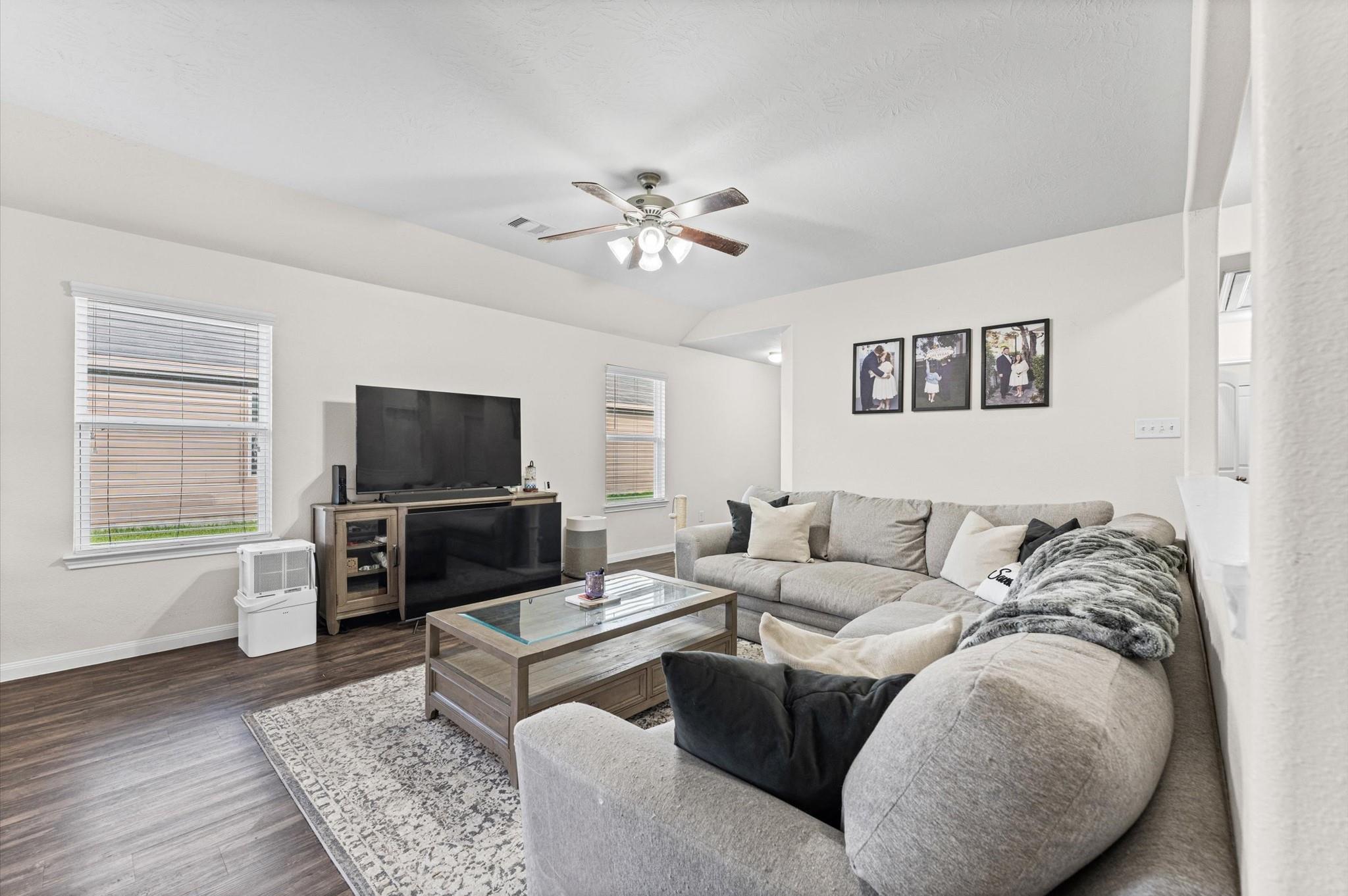 Living room with a gray sectional, glass-top coffee table, and television beneath a ceiling fan.