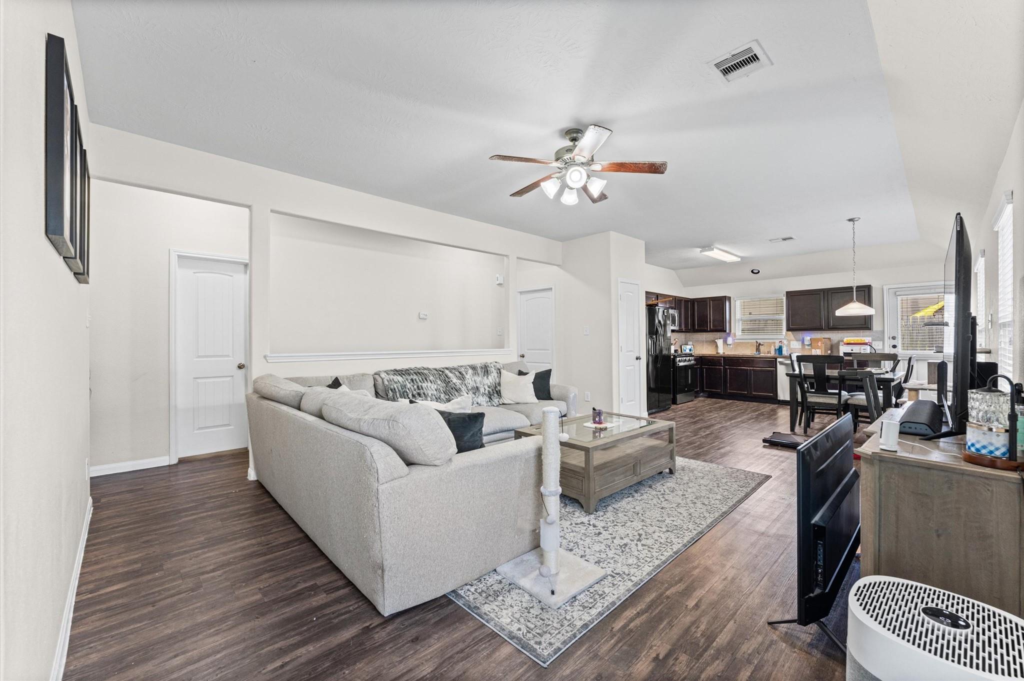 Open-plan living room with a gray sectional, adjoining dark-cabinet kitchen, and wood-look floors.