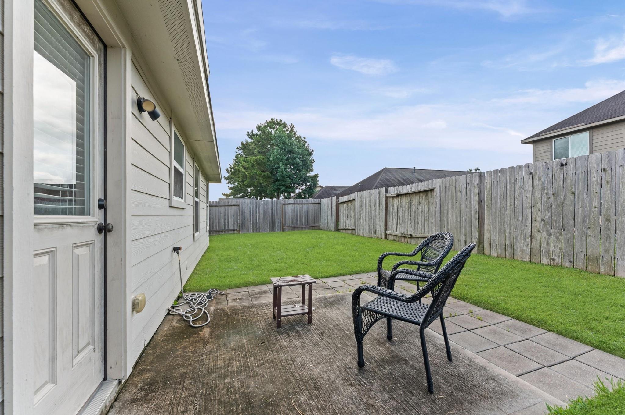 Backyard patio with two wicker chairs and a small table beside a fenced green lawn.