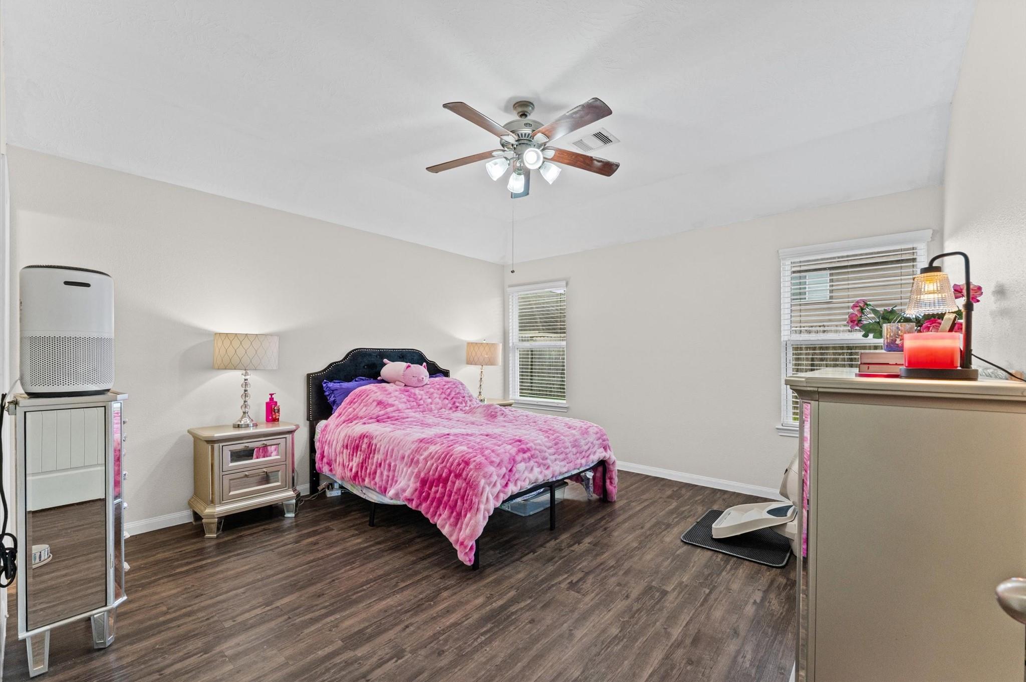 Bedroom with a bright pink bedspread, dark headboard, ceiling fan, and dark wood-look flooring.
