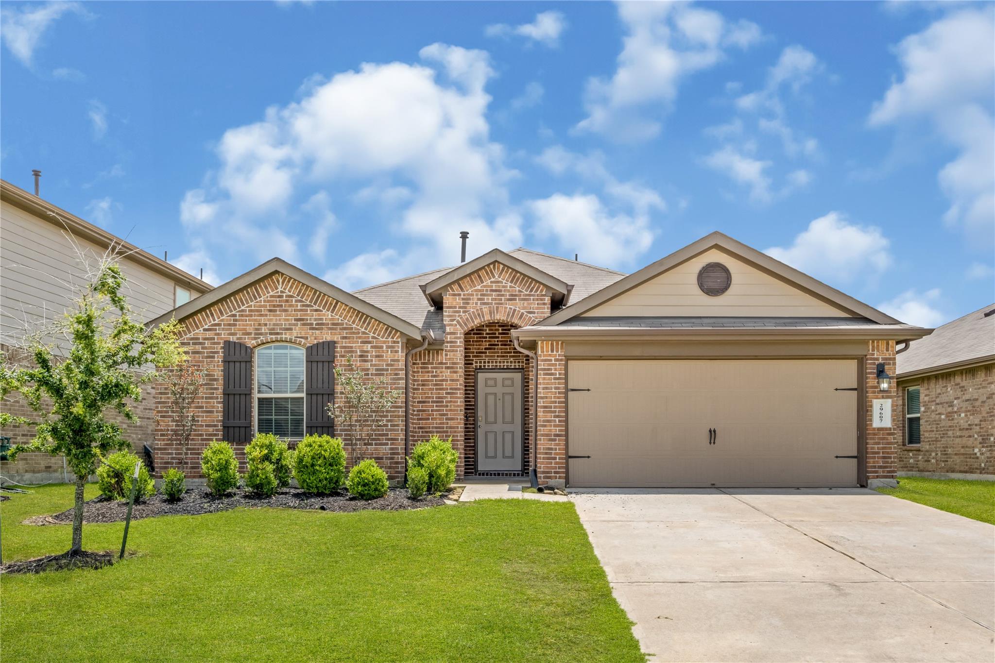 Brick-front house with a tan two-car garage, dark shutters, and a landscaped lawn.