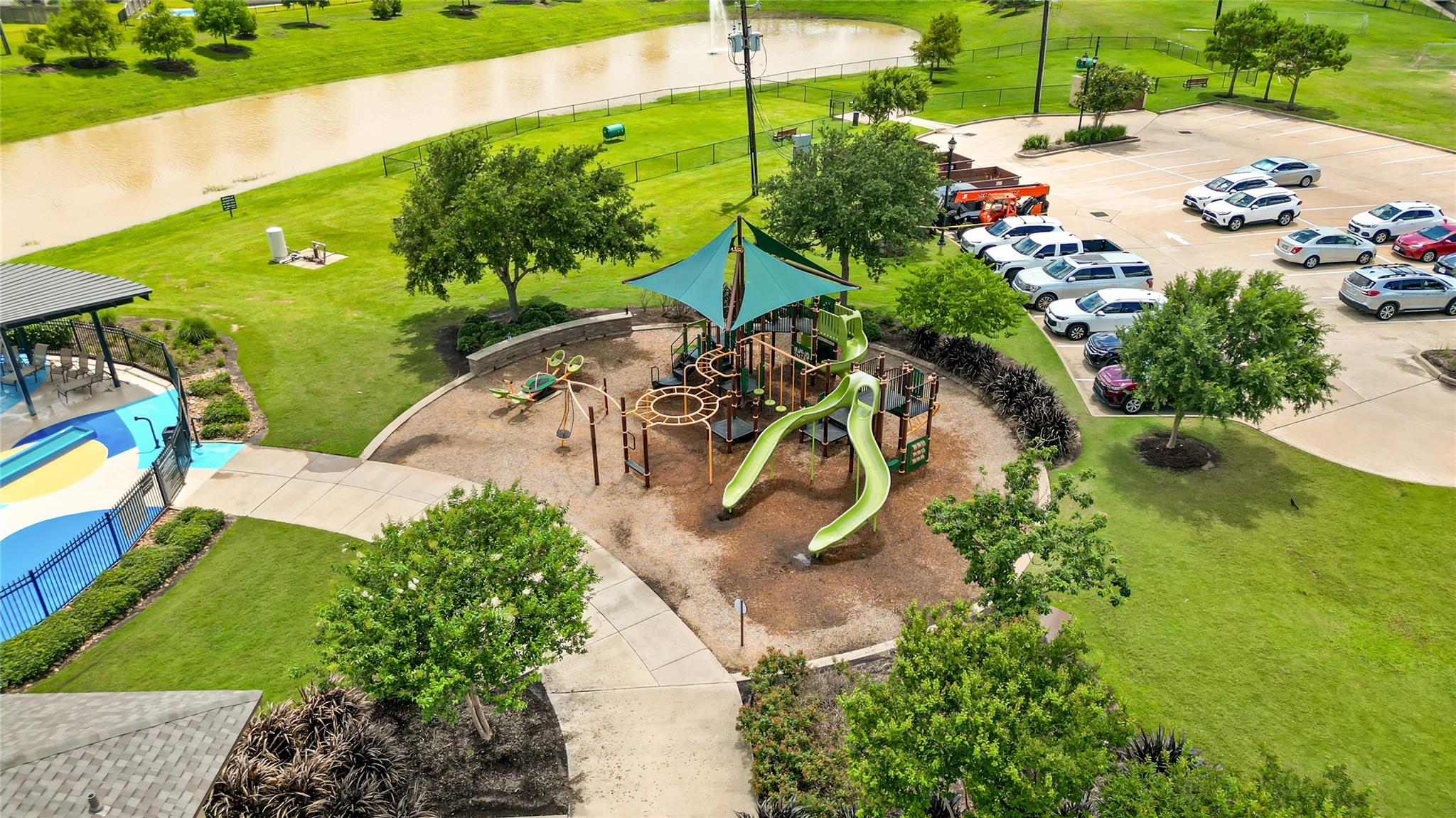 Community playground with shade canopies, swimming pool, parking area, and waterway, seen from above.