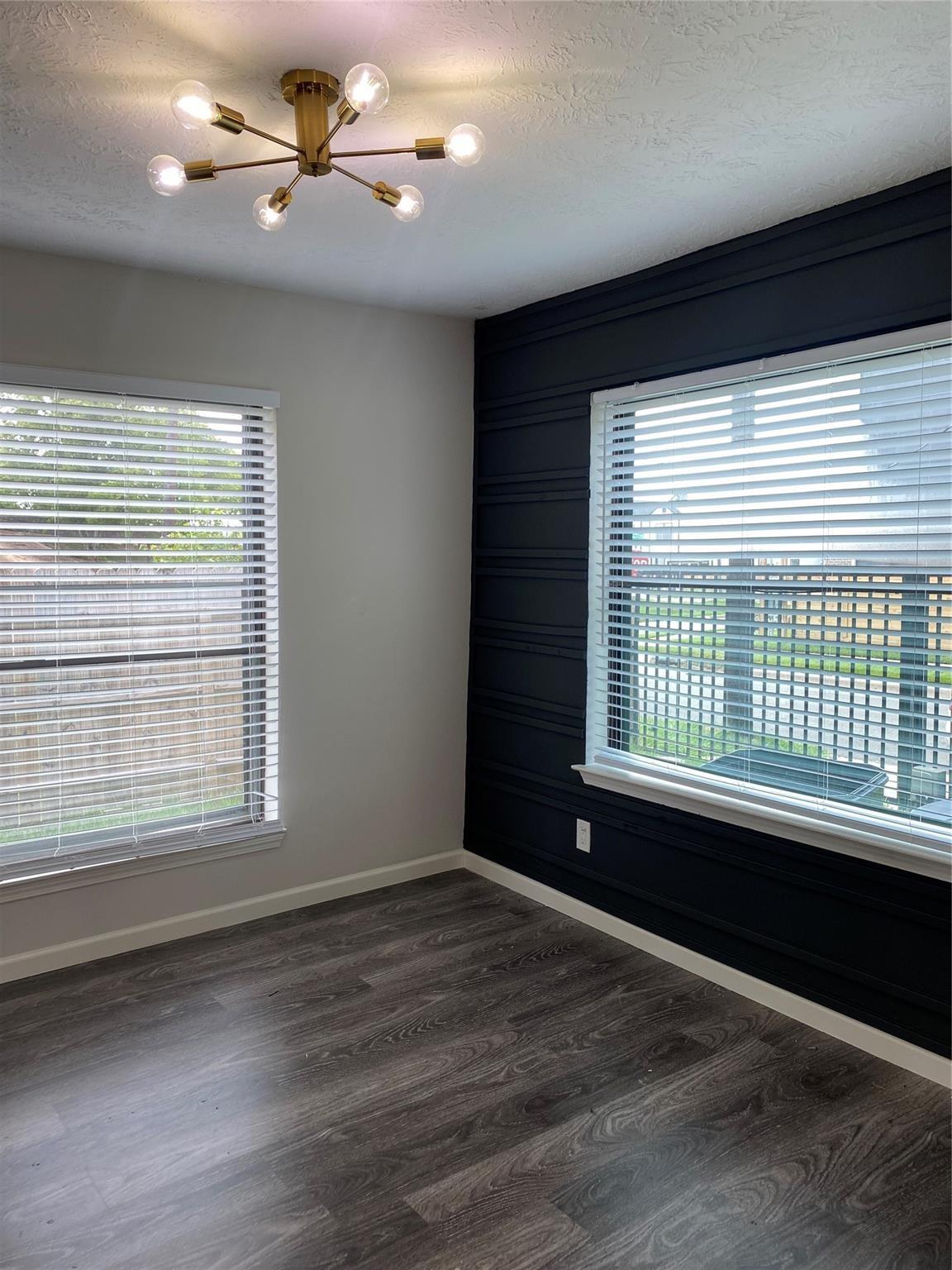 Empty room with a navy paneled accent wall, two blinded windows, dark flooring, and a brass ceiling light.