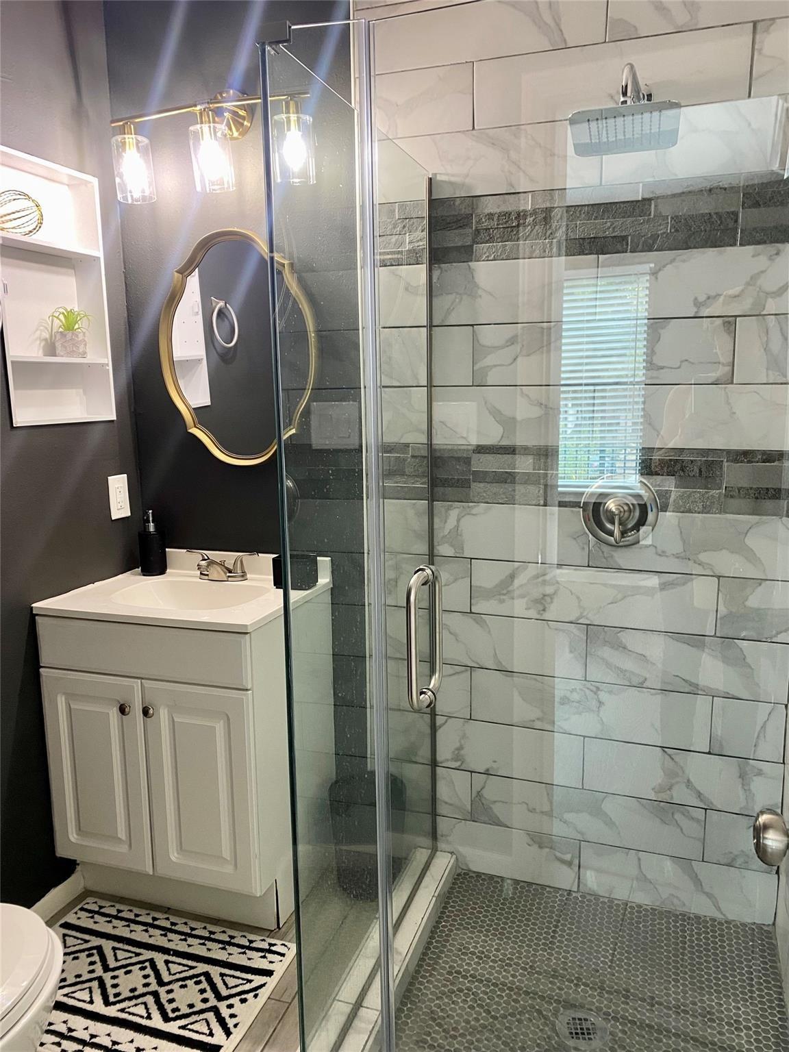 Bathroom with a glass walk-in shower, marble-look tile, and white vanity beneath a gold-framed mirror.