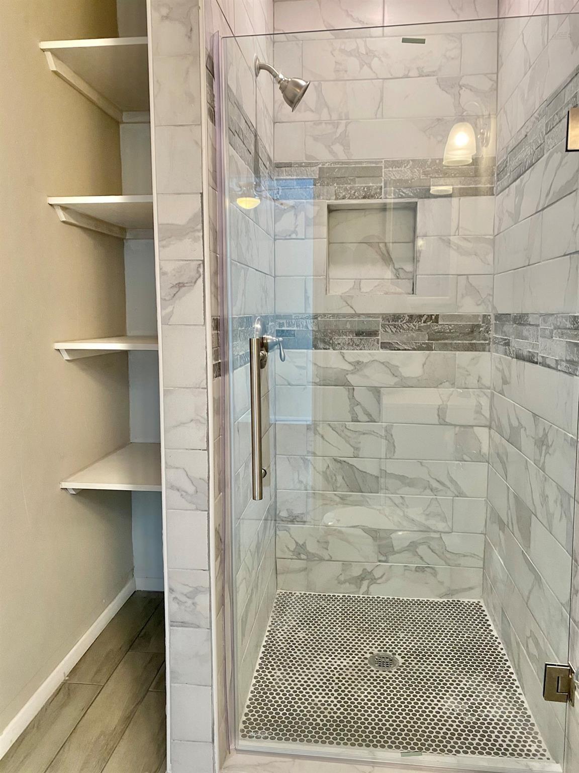 Walk-in shower with marble-look tile, gray accents, a recessed niche, and open shelving beside it.