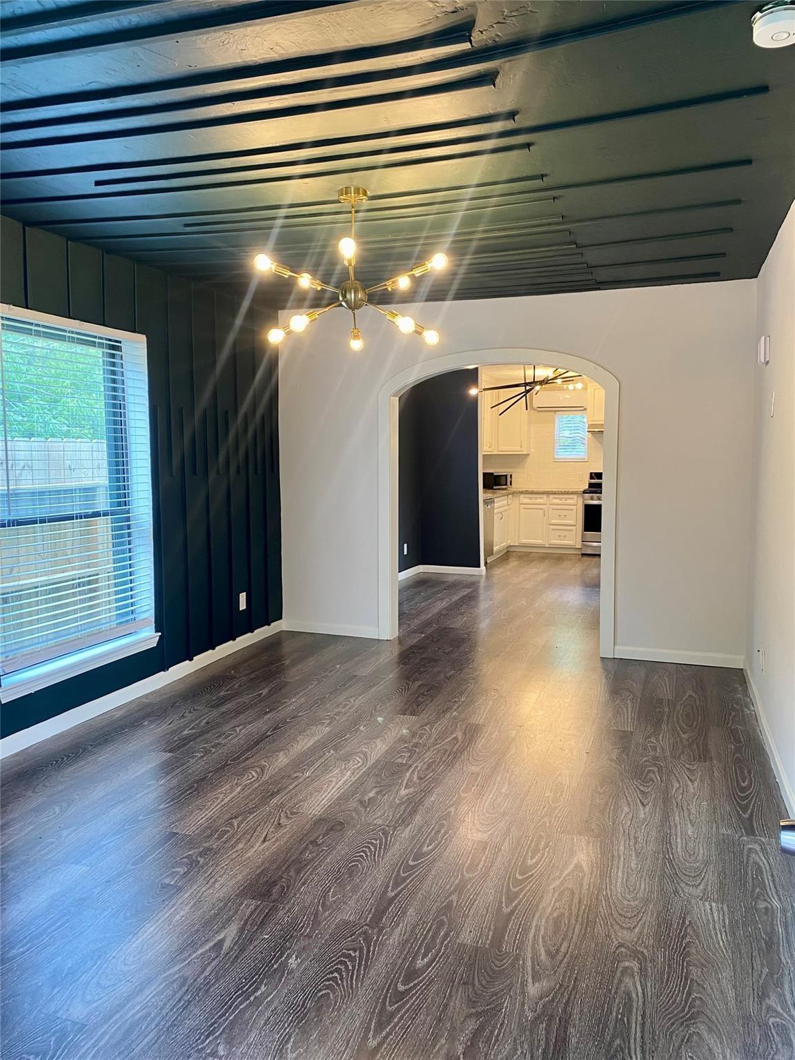 Empty room with dark wood-look floors, teal accent wall, exposed beams, and an arched opening to the kitchen.