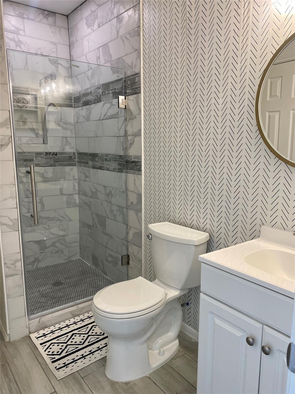 Bathroom with a marble-look tiled walk-in shower, patterned wall, white vanity, and round mirror.
