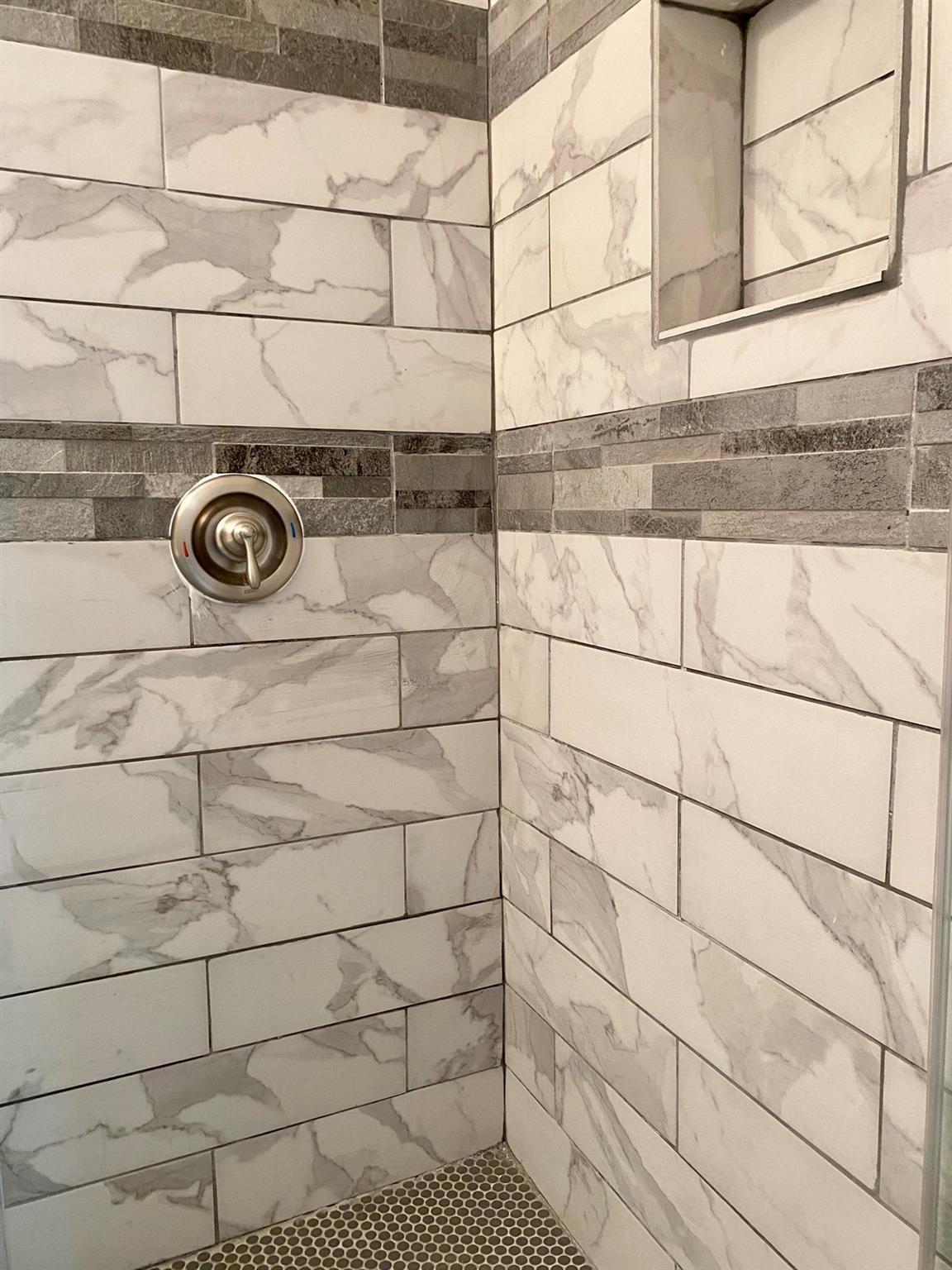 Tiled shower with marble-look walls, gray stone accent band, and hexagonal mosaic floor.