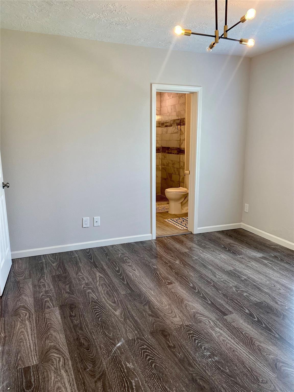 Empty room with dark wood-look flooring and a chandelier, opening to a tiled bathroom.