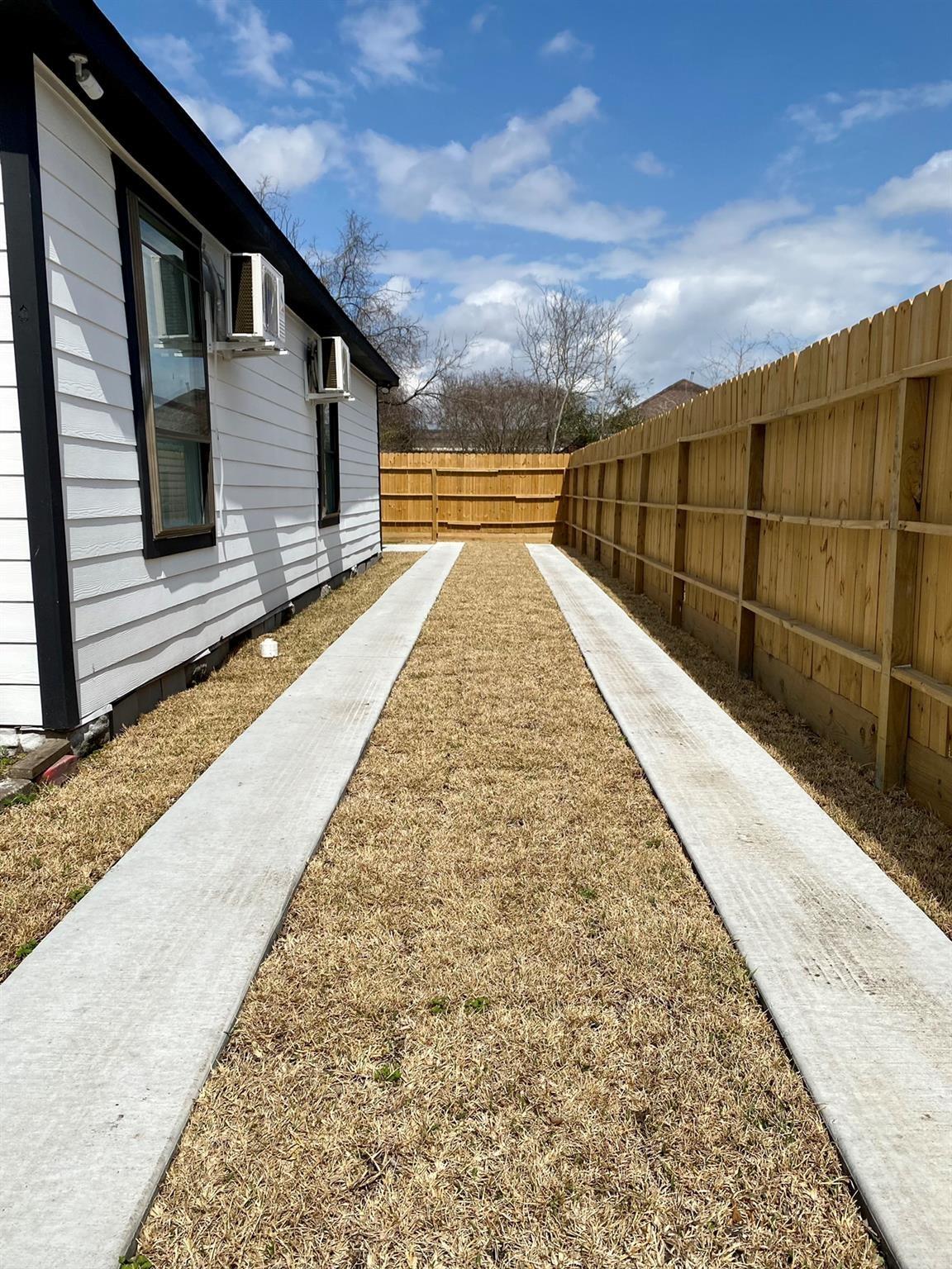 Enclosed side yard with two concrete strips through dormant grass beside a white-sided house.