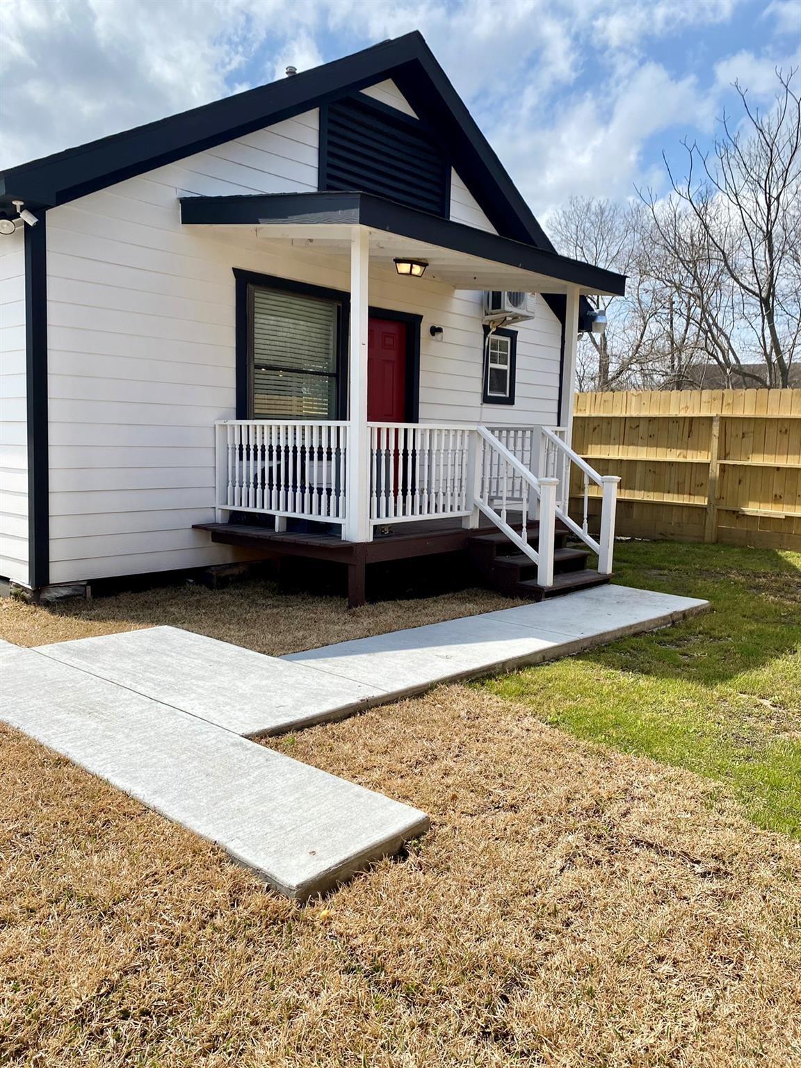 White house with black trim, a red door, and a covered porch beside a concrete walkway.