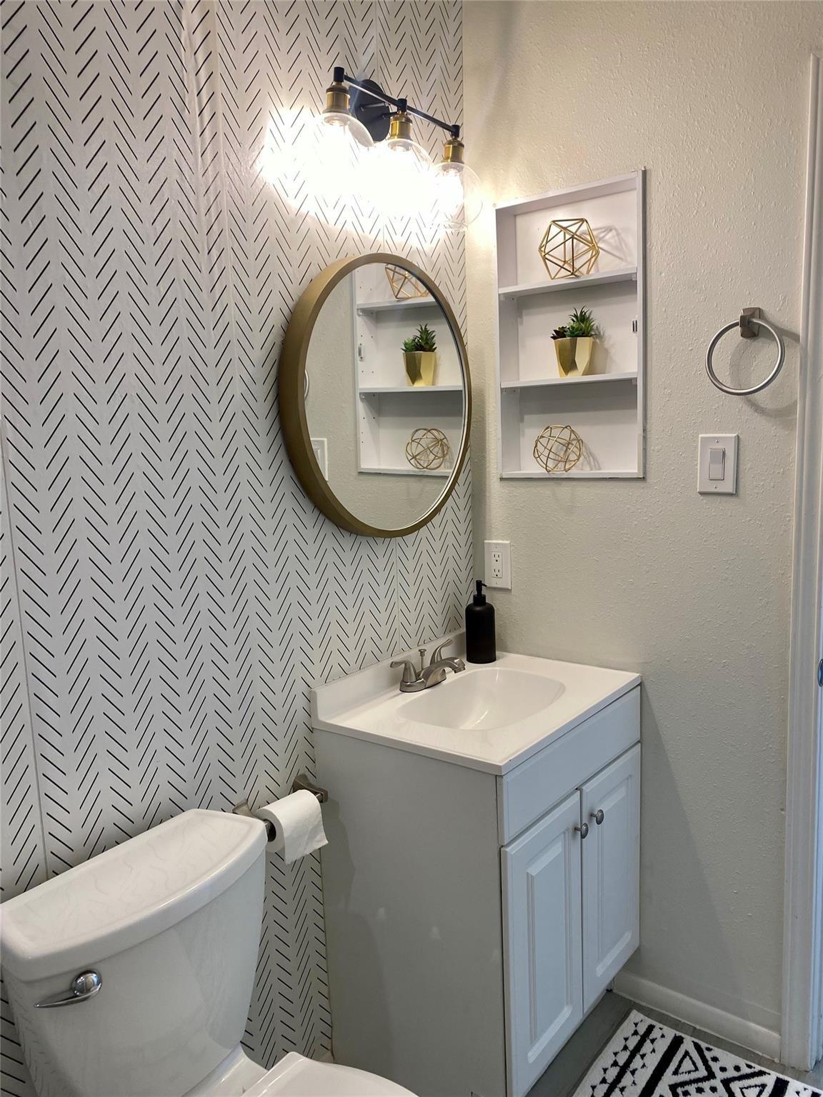 Bathroom with white vanity, round gold-framed mirror, and black-and-white patterned accent wall.
