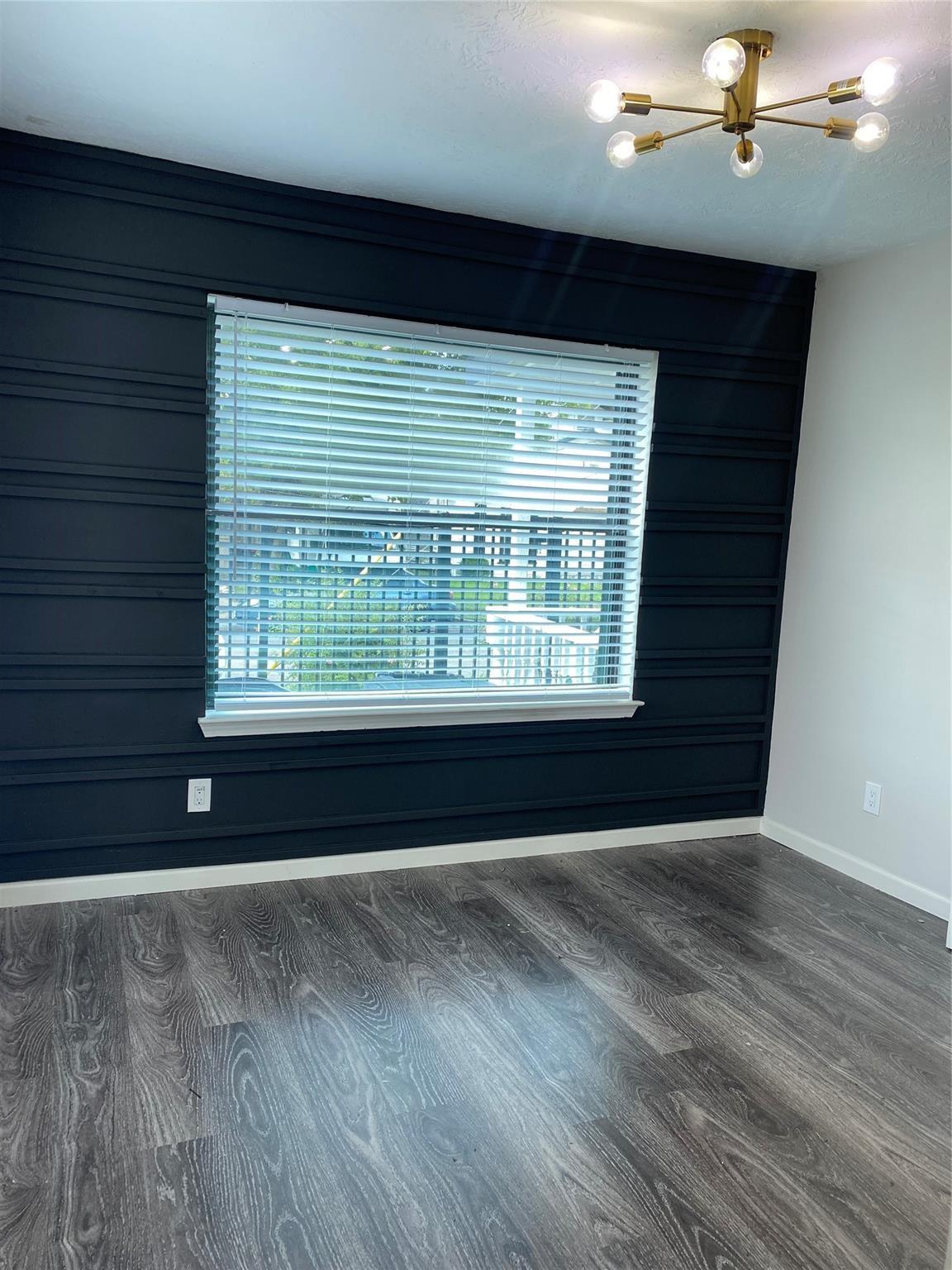 Empty room with a navy paneled accent wall, wide blinded window, gray plank flooring, and brass ceiling light.