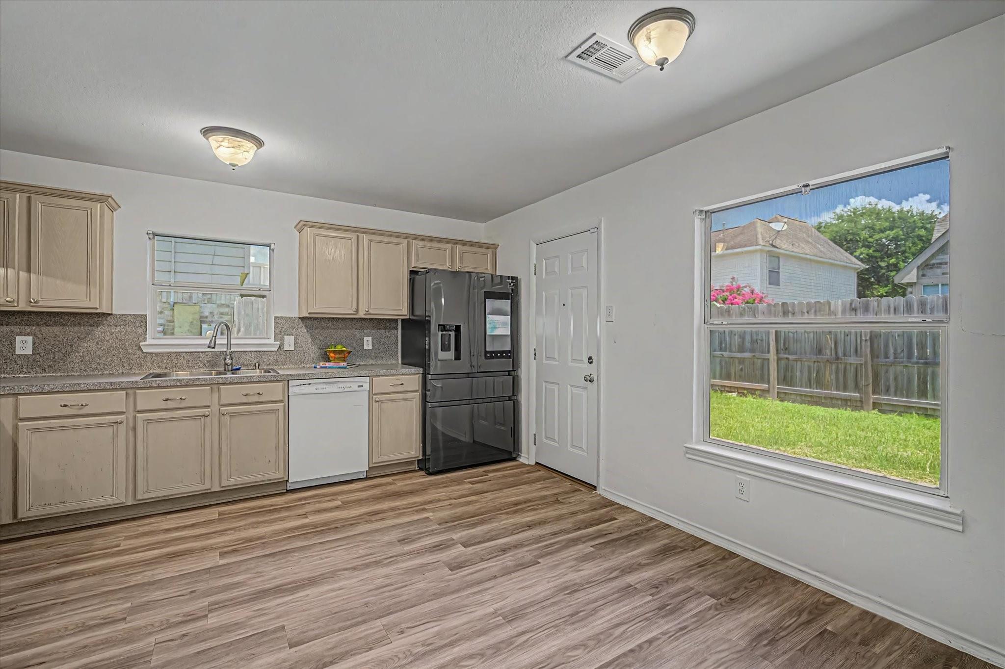 Kitchen with light wood cabinets, black refrigerator, and a large window overlooking a fenced backyard.