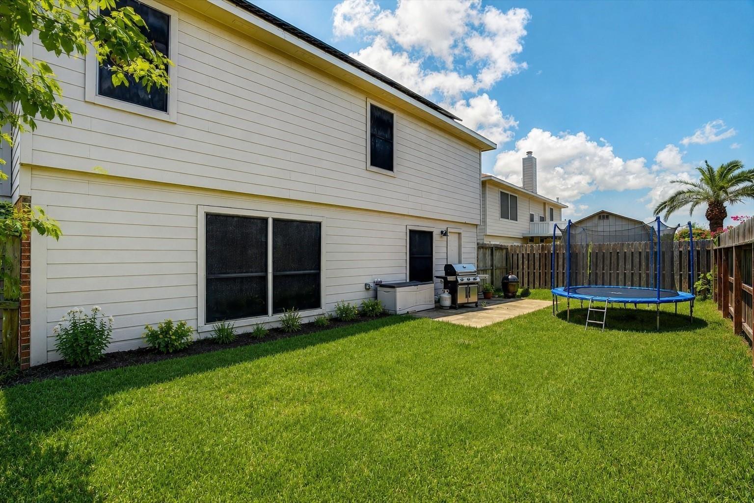 Rear of a two-story house beside a fenced lawn with a trampoline and outdoor grill.