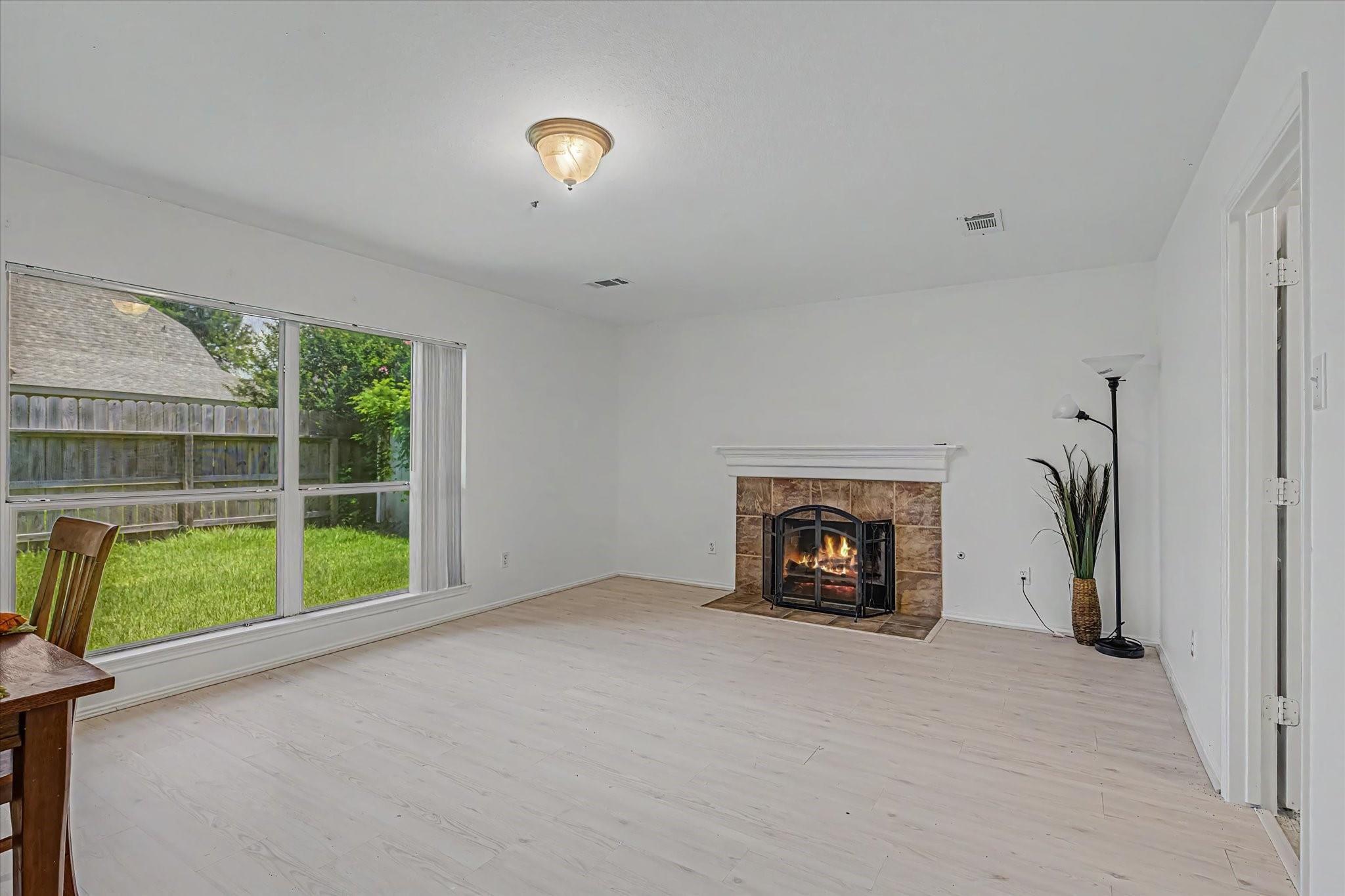 Living room with a lit stone fireplace and large windows overlooking a fenced backyard.