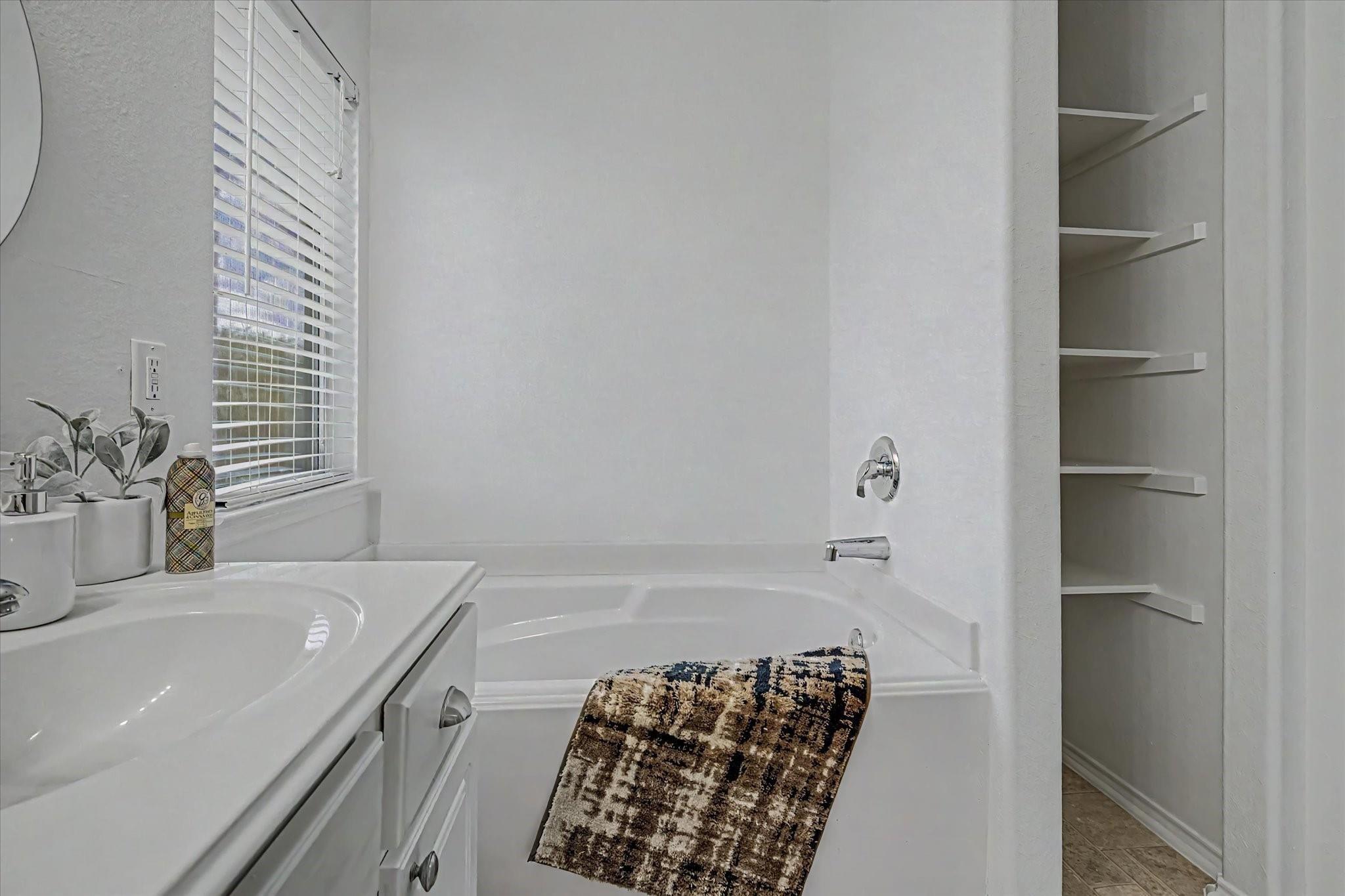 Bathroom with a white bathtub, vanity, window blinds, and built-in shelves.