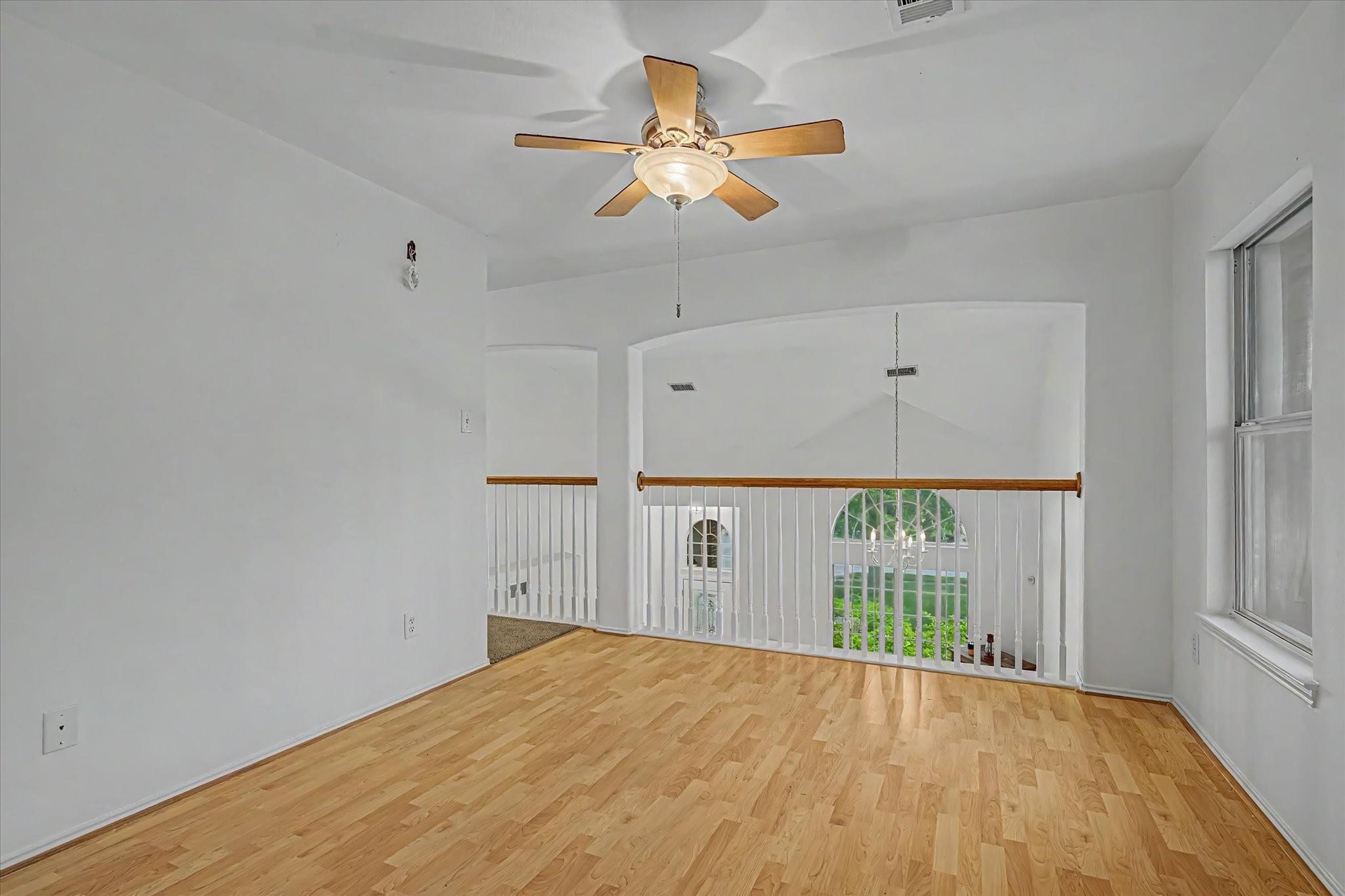 Empty upstairs landing with hardwood floors, ceiling fan, and railing overlooking a bright two-story foyer.