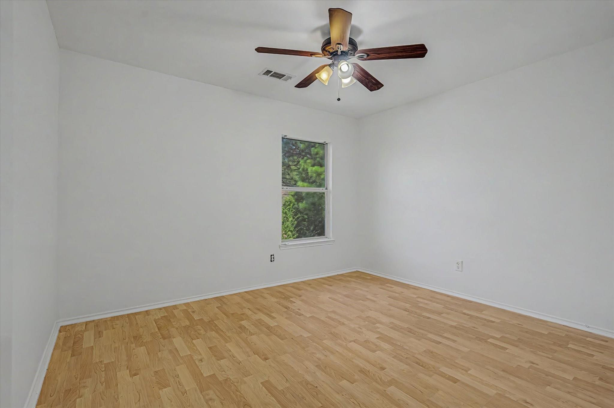 Vacant room with hardwood floors, a window overlooking greenery, and a ceiling fan with lights.