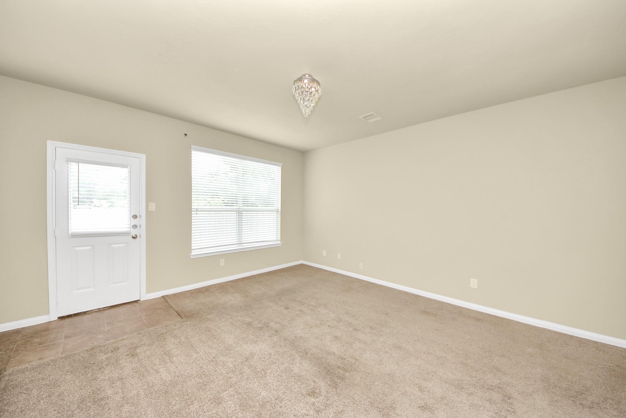 Empty carpeted room with a tiled entry, white door, window blinds, and hanging ceiling light.