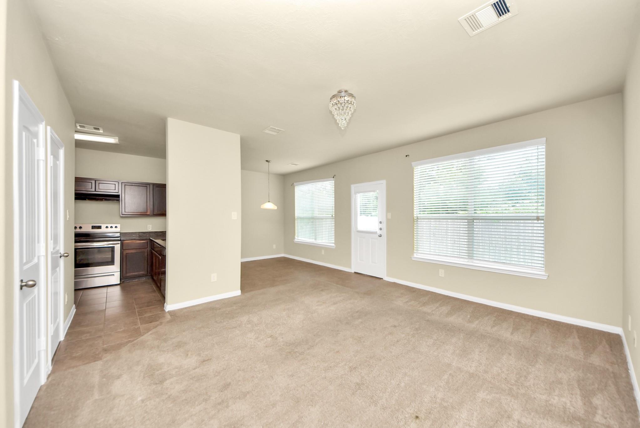 Vacant living and dining area with beige carpet, crystal chandelier, and kitchen visible beyond.