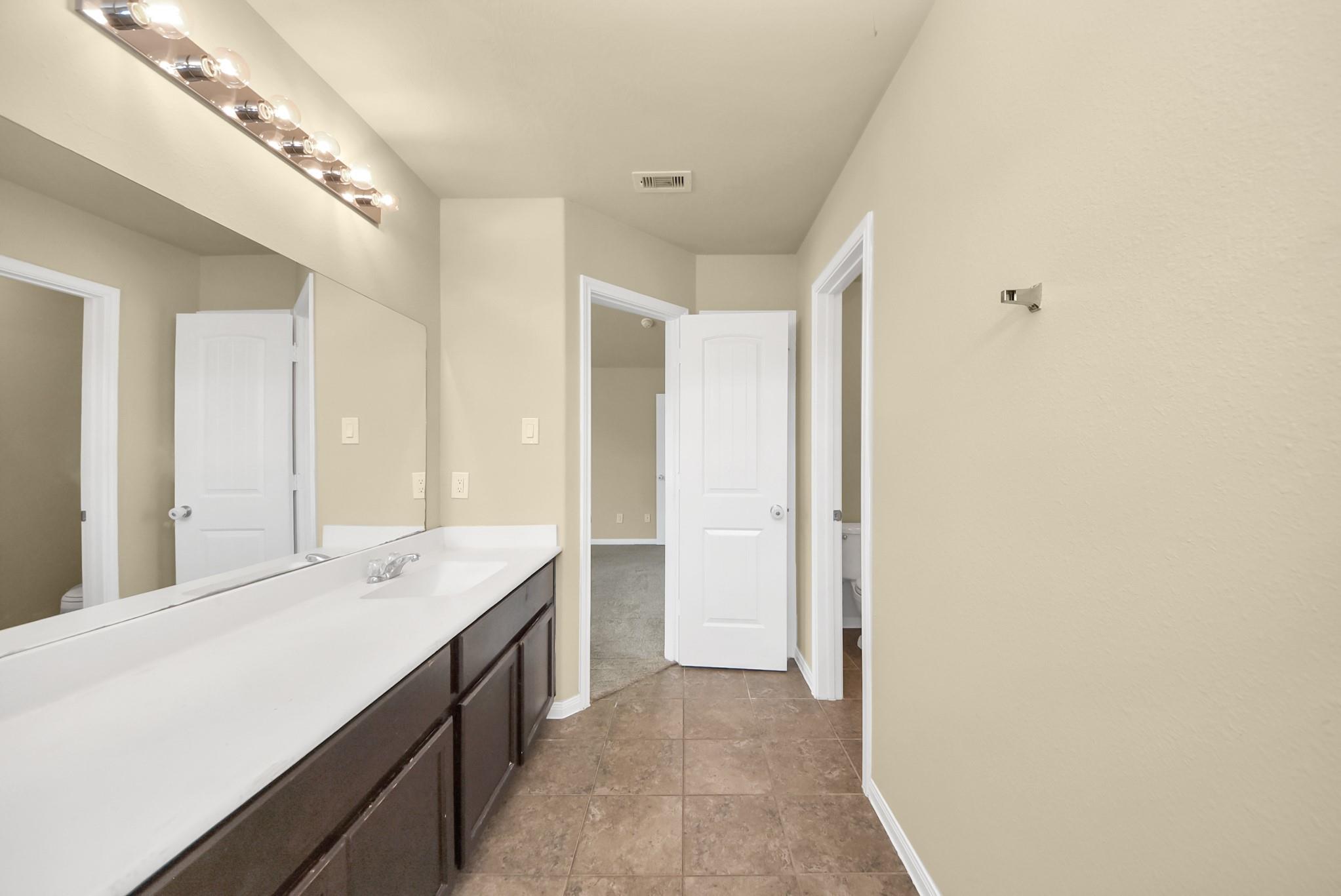 Bathroom vanity with a white countertop, dark cabinets, large mirror, and brown tile floor.