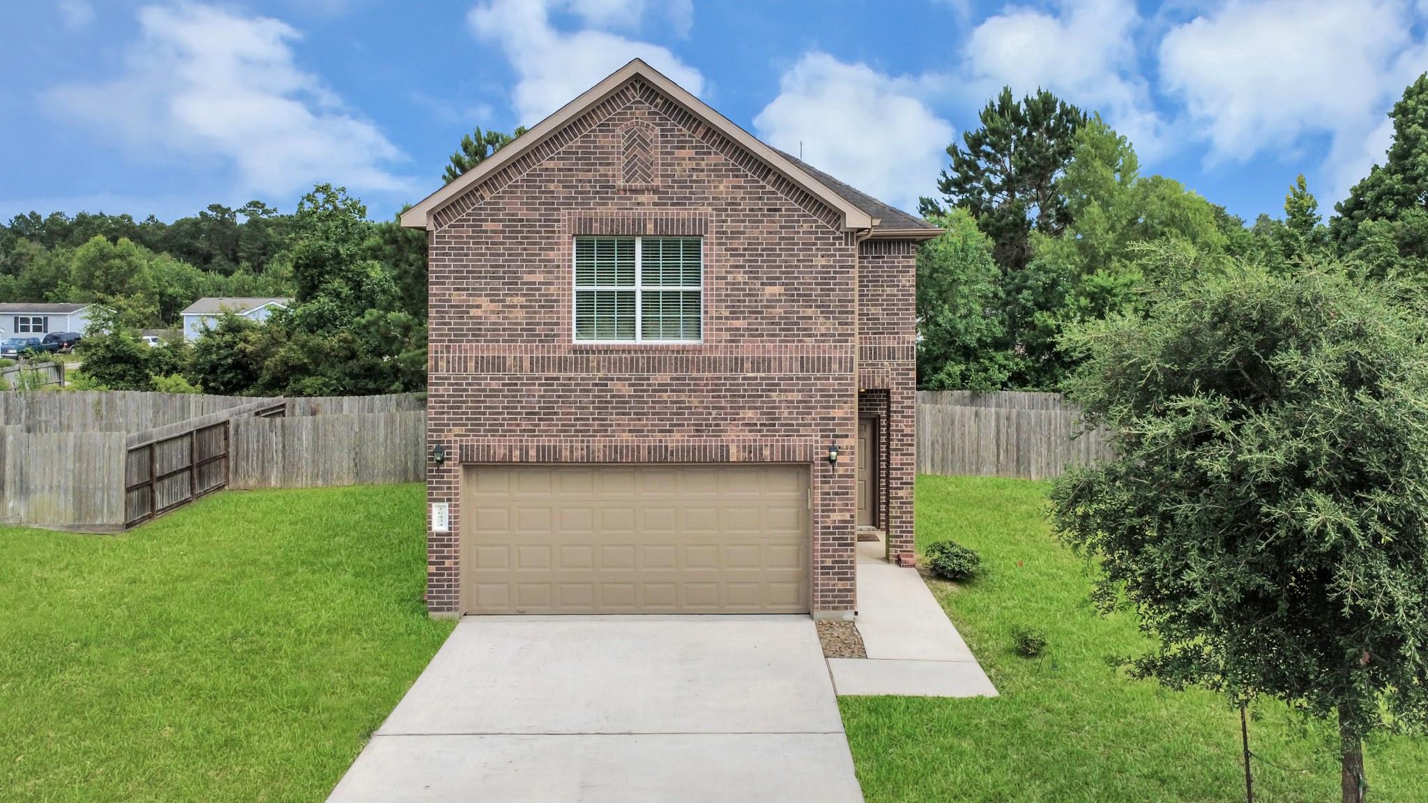 Two-story brick house rear with a double garage, concrete driveway, fenced lawn, and mature trees.