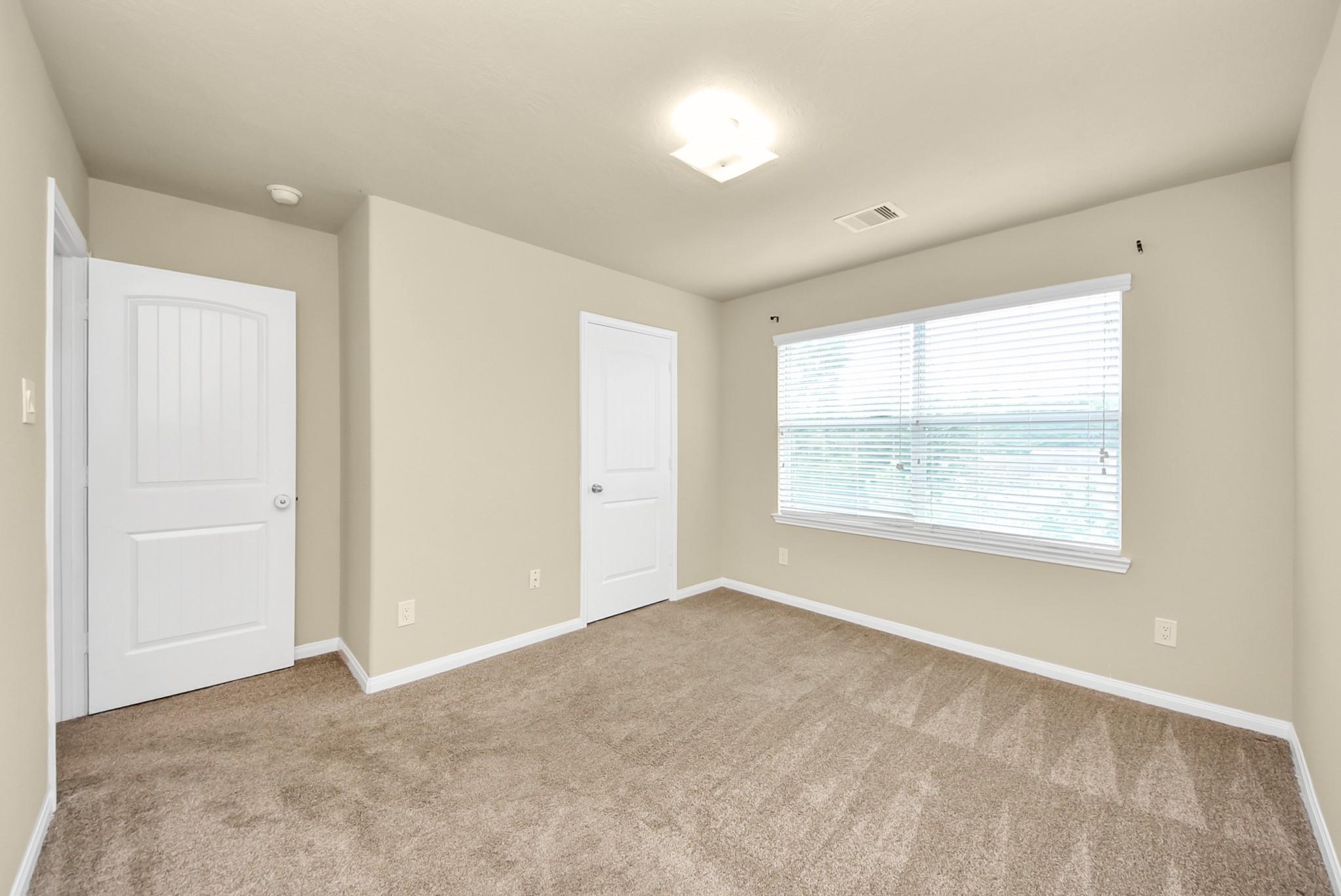 Empty bedroom with tan carpeting, beige walls, and a wide window with white blinds.