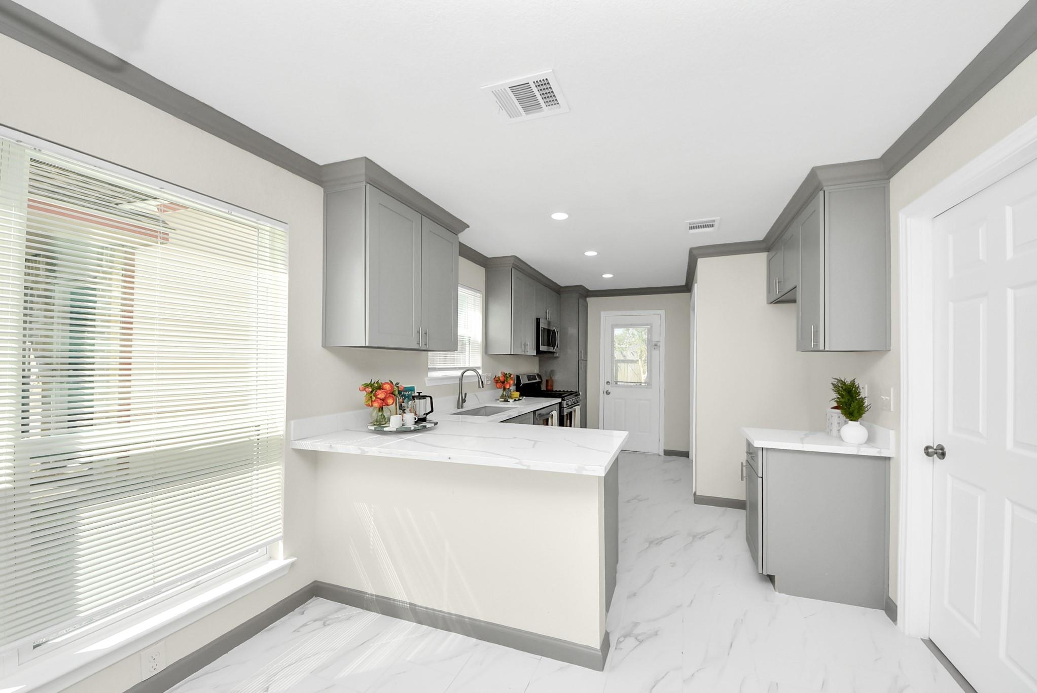 Bright kitchen with gray cabinets, white peninsula, marble-look flooring, and a large window with blinds.